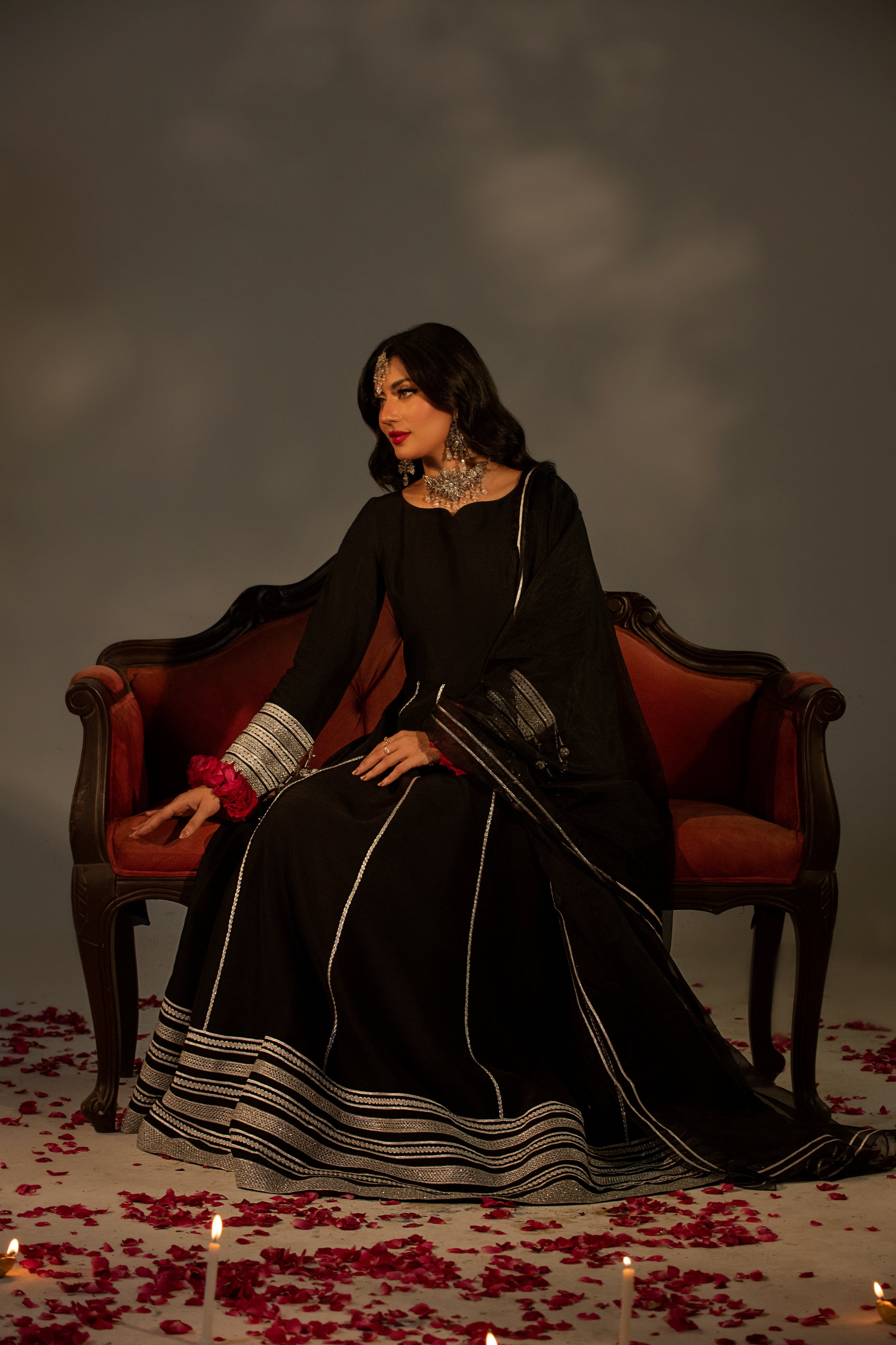 Woman in a black dress sitting on a red couch with candles and rose petals on the floor.
