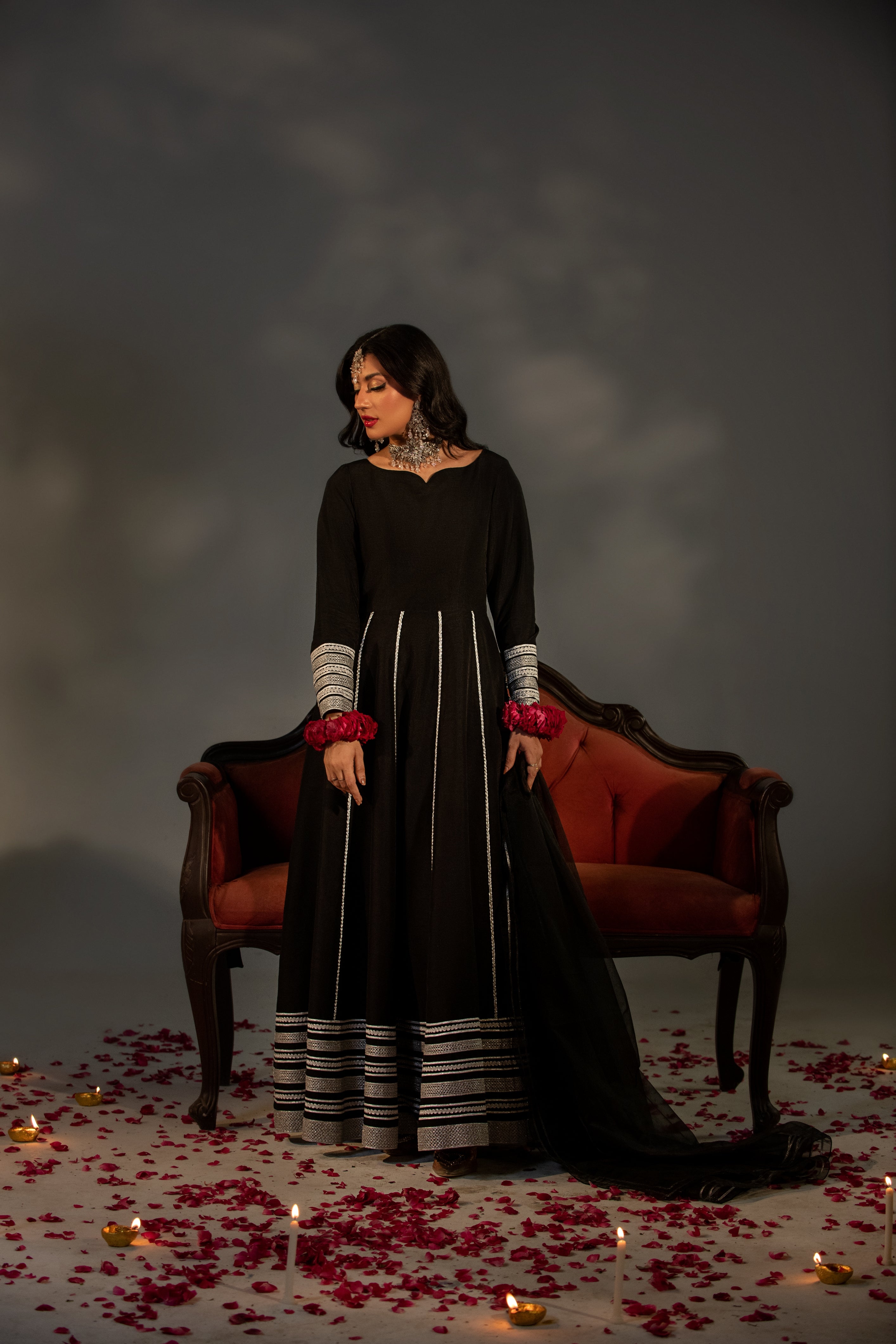Woman in a black dress with red accents standing in front of a dark background with candles and rose petals.