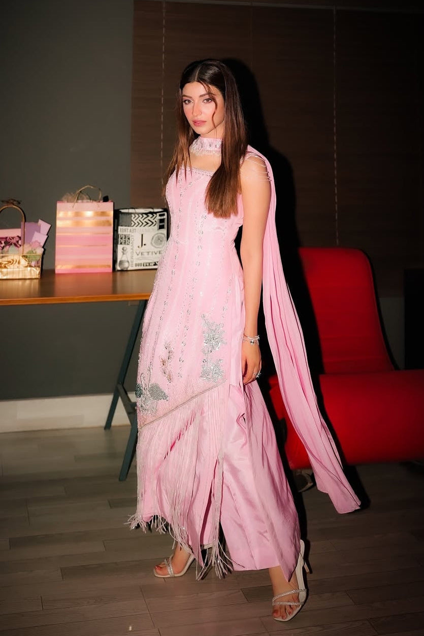 Woman in a pink saree standing indoors with a red chair and wooden table in the background.