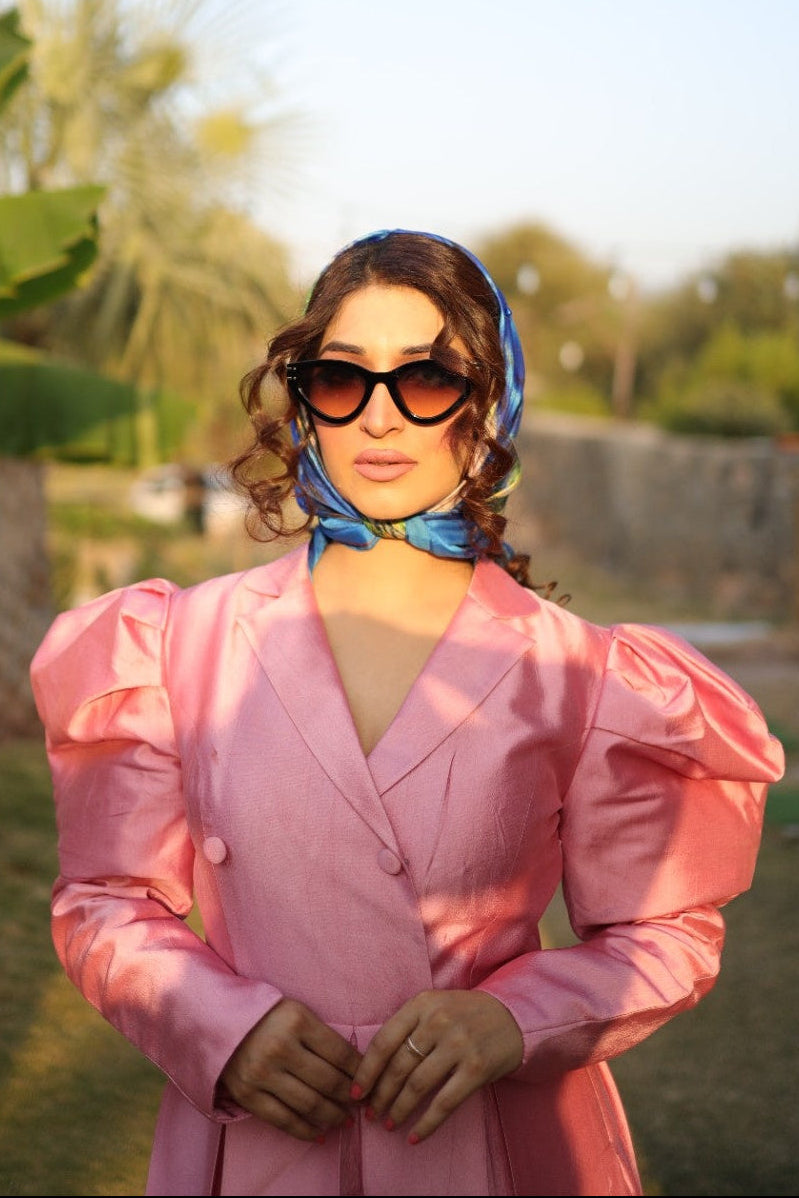 Woman in a pink outfit with sunglasses and a headscarf standing outdoors.