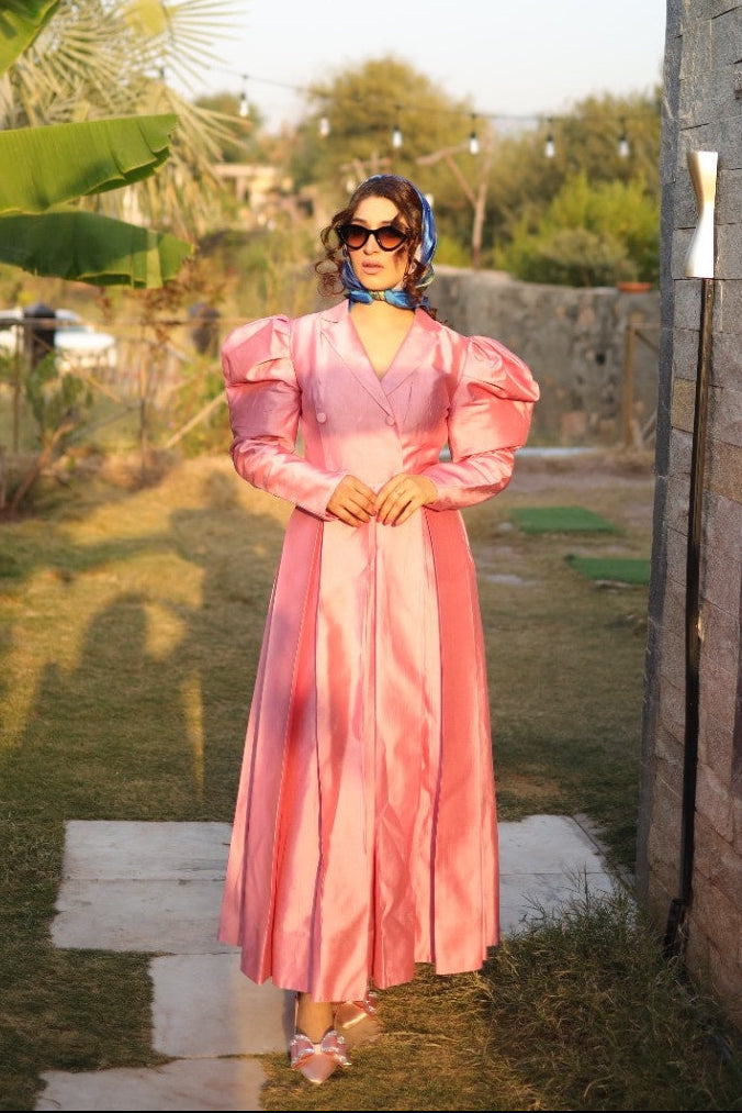 Woman in a pink dress standing outdoors with greenery and a building in the background