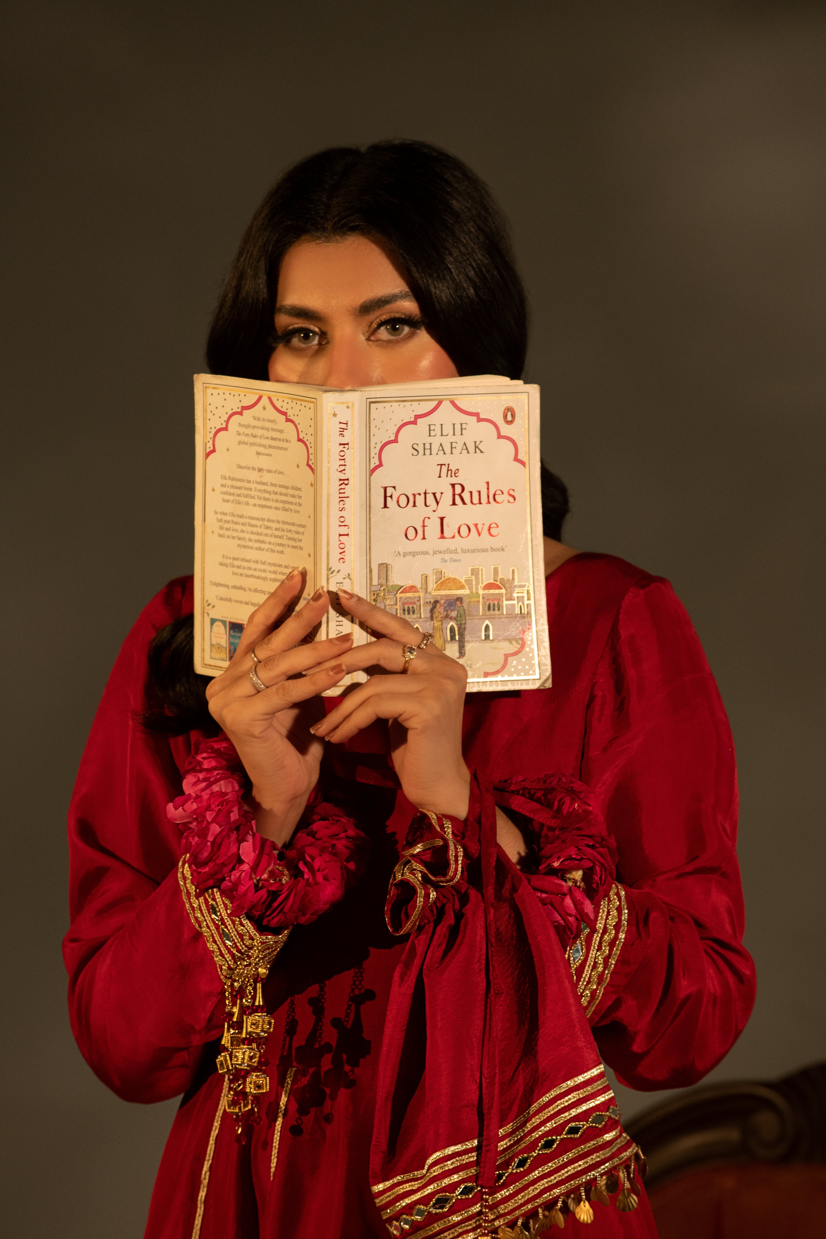 Woman in traditional attire holding a book titled 'Forty Rules of Love' by Elif Shafak against a dark background.