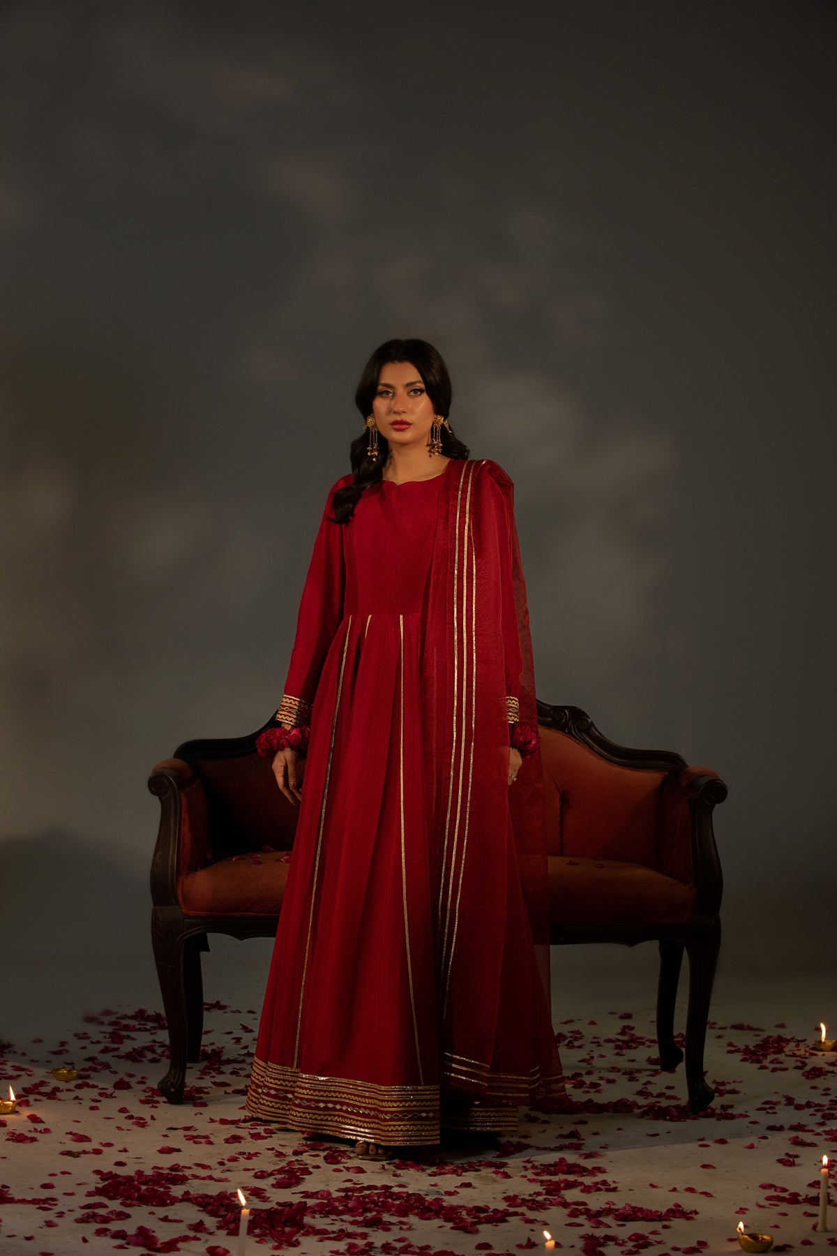 Woman in a red traditional outfit standing in front of a dark background with candles and rose petals.