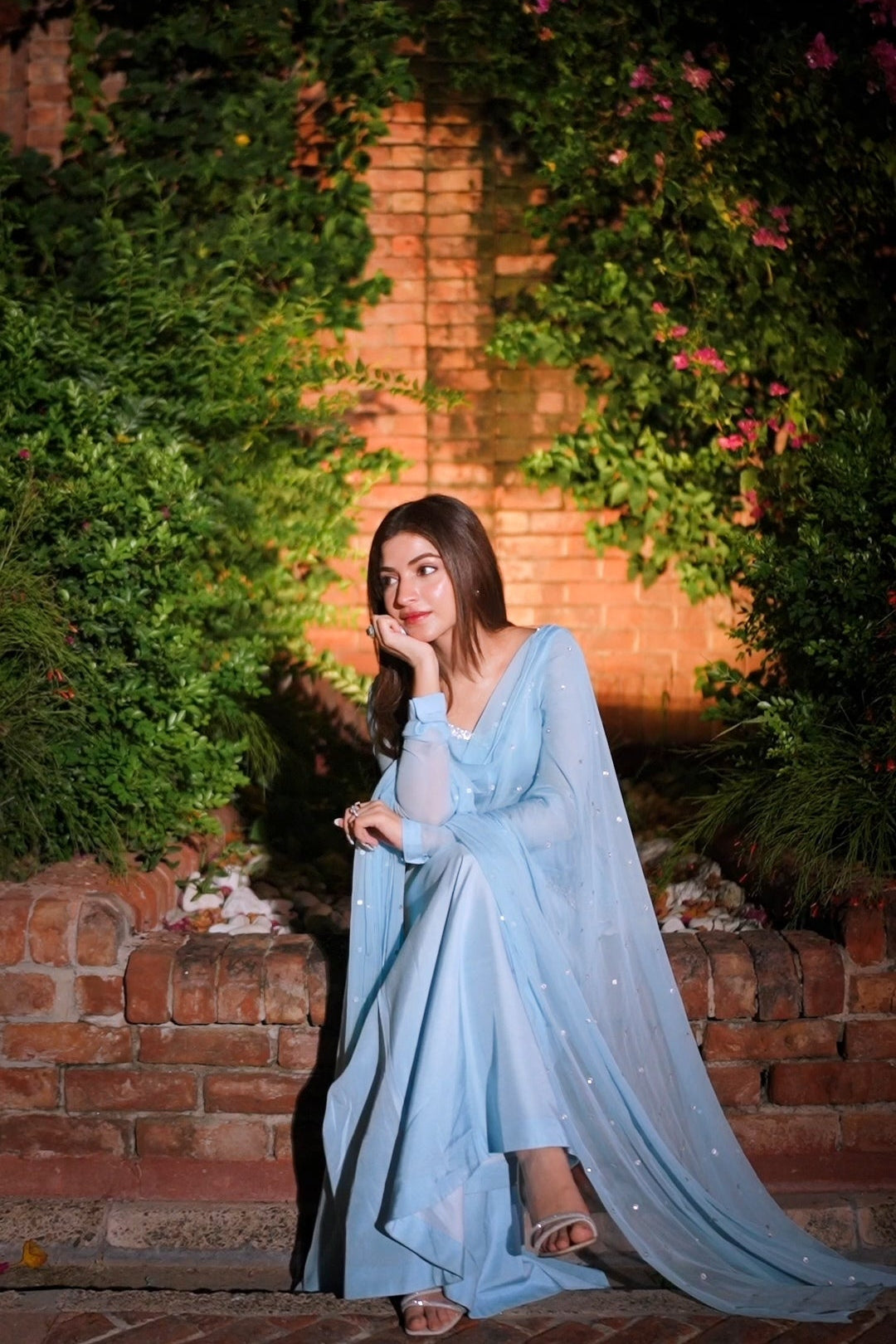 A woman wearing a flowing light blue traditional dress with a sheer dupatta sits on a brick ledge in front of a brick wall covered in green ivy and pink flowers at night.