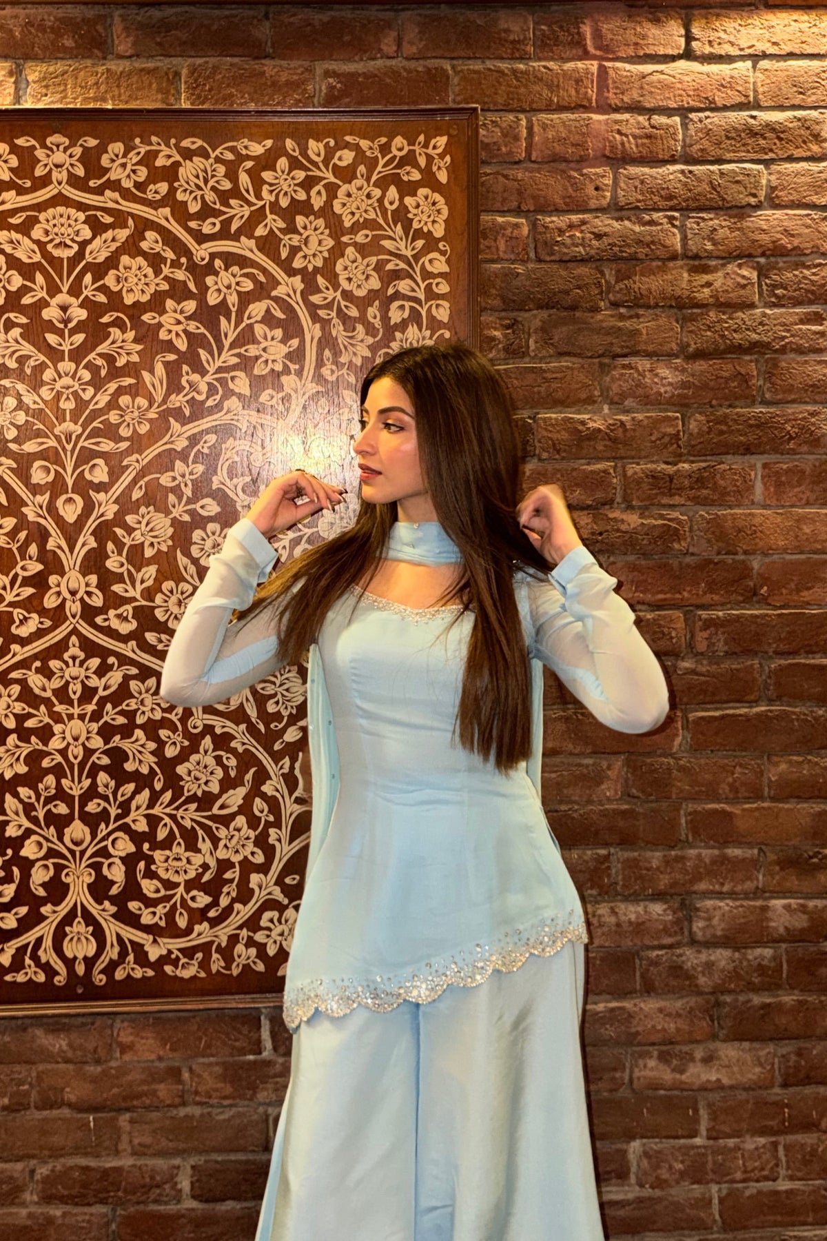 A woman modeling a light blue traditional outfit with sheer long sleeves and lace trim, standing in front of a brown patterned wall panel and brick wall.