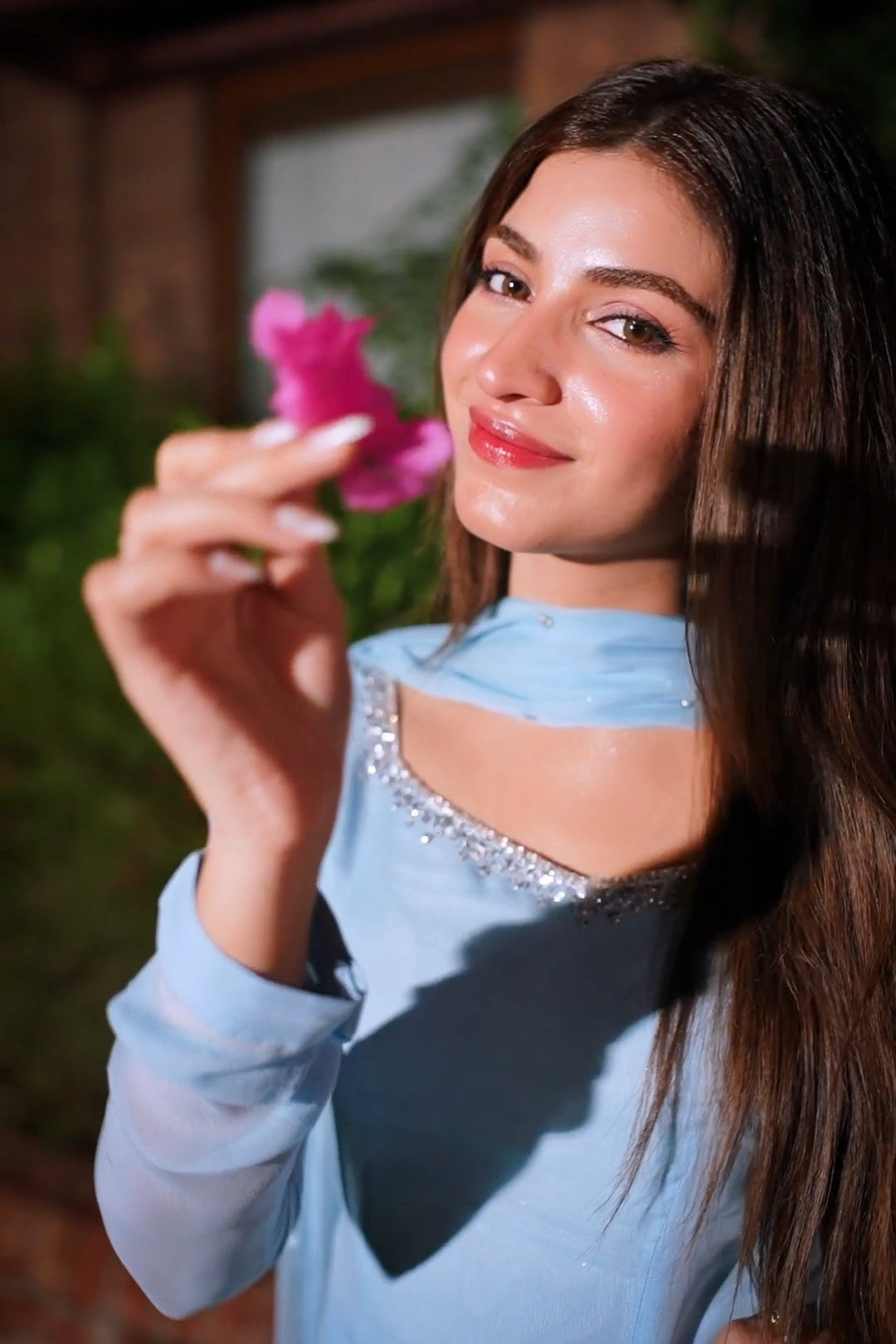 A woman with long dark hair wearing a light blue sleeveless top with silver embellishments, holding a pink flower in her hand against a softly blurred outdoor background.