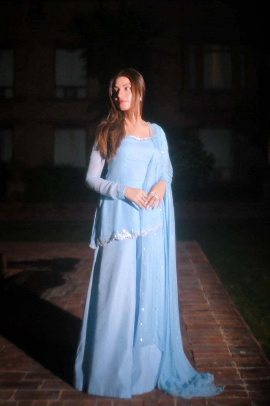 A woman wearing a light blue traditional outfit with a long draped shawl, standing on a brick path at night in front of a dark building.