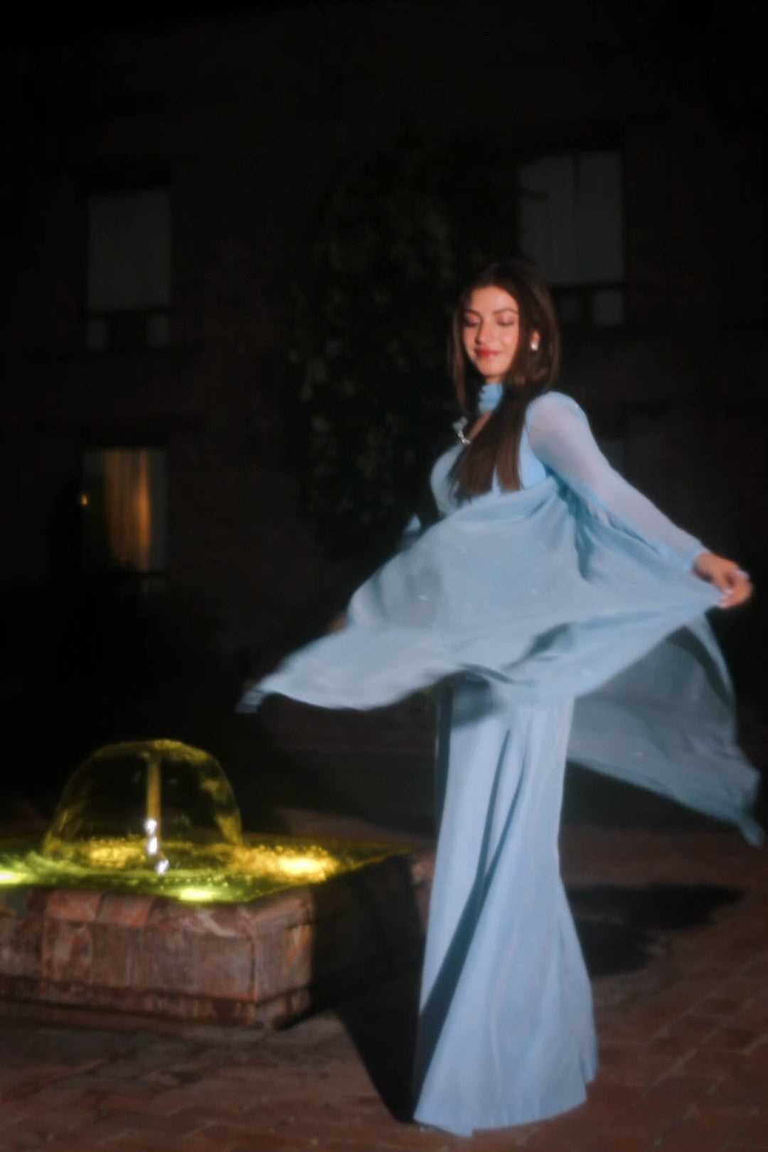 A woman in a flowing light blue long-sleeve maxi dress twirls outdoors at night beside a lit fountain, with a dark building in the background.