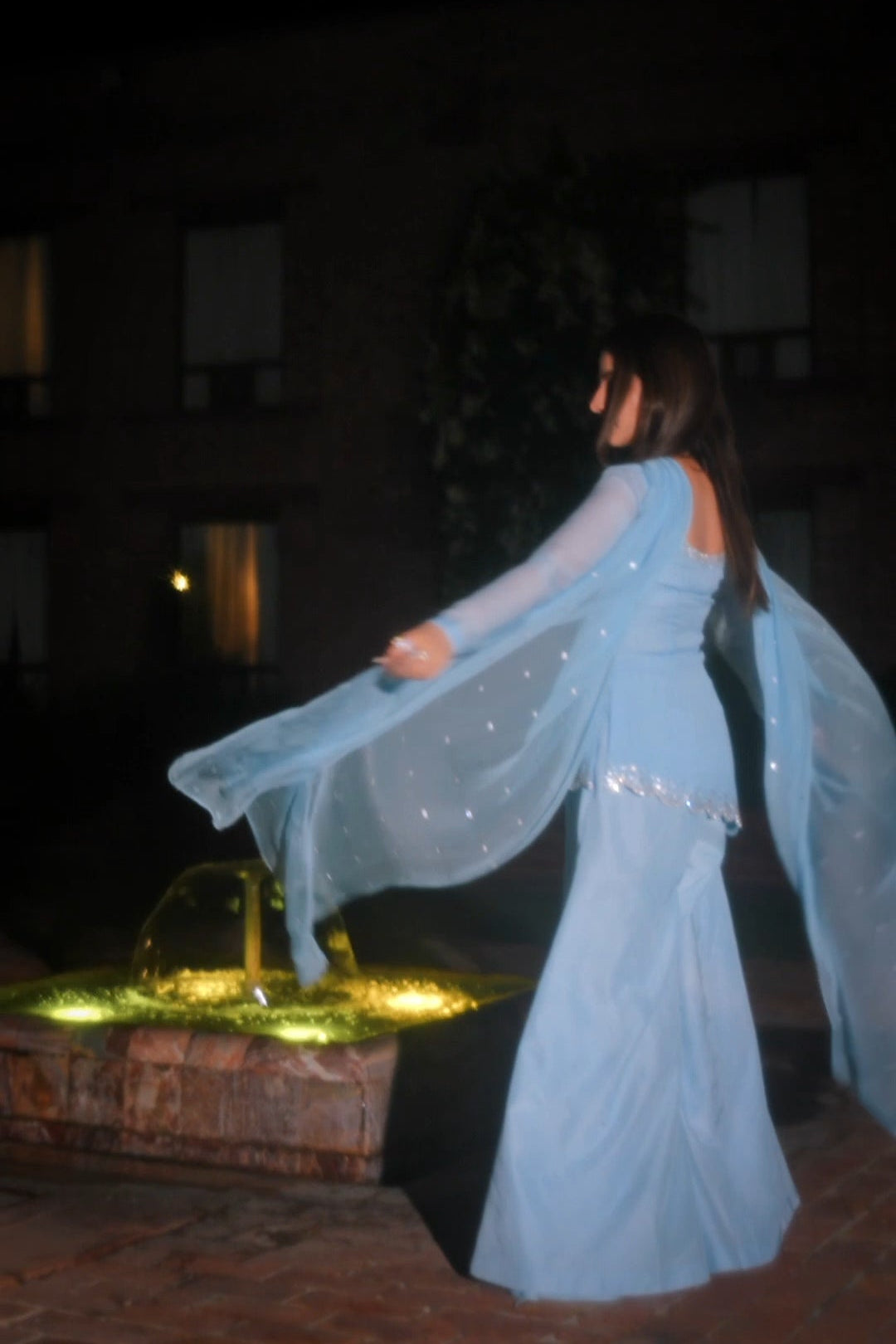 A woman in a flowing light blue embellished dress twirling outdoors at night near a lit fountain, with a dark building in the background.