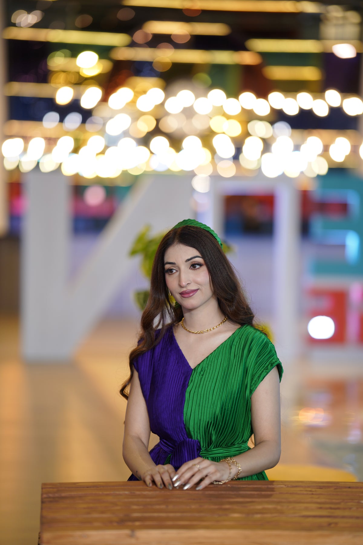 A woman wearing a pleated two-tone dress in purple and green, sitting behind a wooden table in a brightly lit indoor setting with blurred lights in the background.