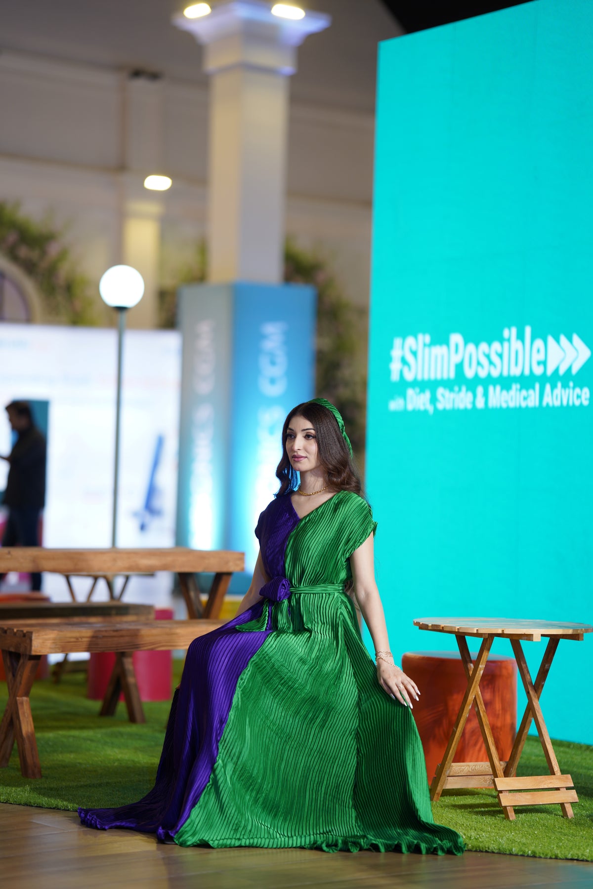 A woman wearing a floor-length pleated dress with a split green and purple color-block design sits on a green carpeted runway. A large teal backdrop on the right displays the text '#SlimPossible with Diet, Stride & Medical Advice'.