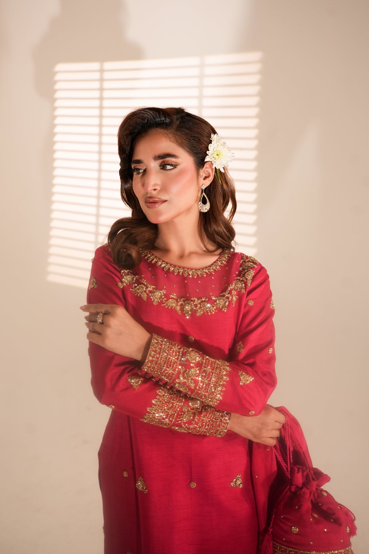 A woman models a vibrant red traditional outfit with intricate gold embroidery on the neckline and cuffs, holding a matching red embroidered clutch bag against a warm beige wall with window blinds casting soft shadows.