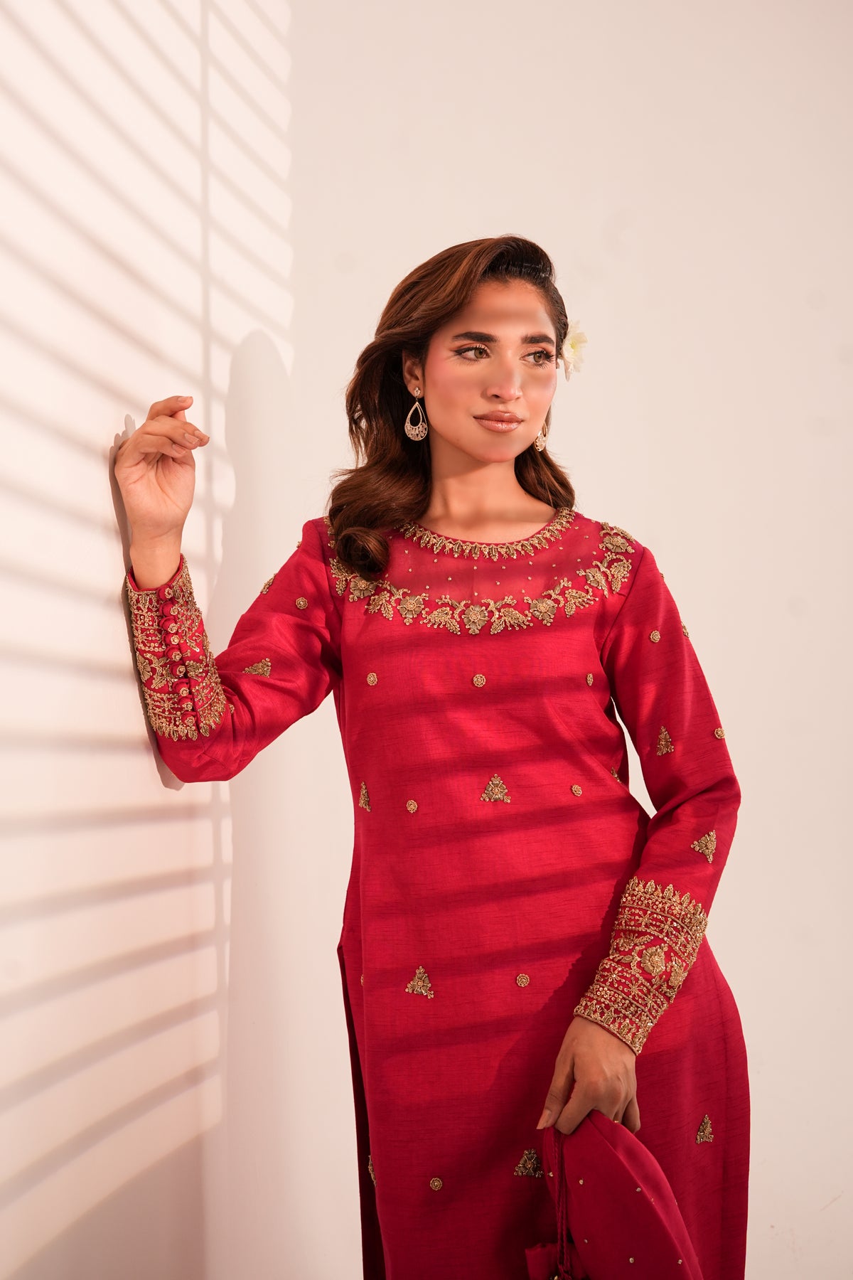 A woman wearing a red long kurta with gold embroidery on the neckline and cuffs, standing against a white wall with window blinds casting shadows.