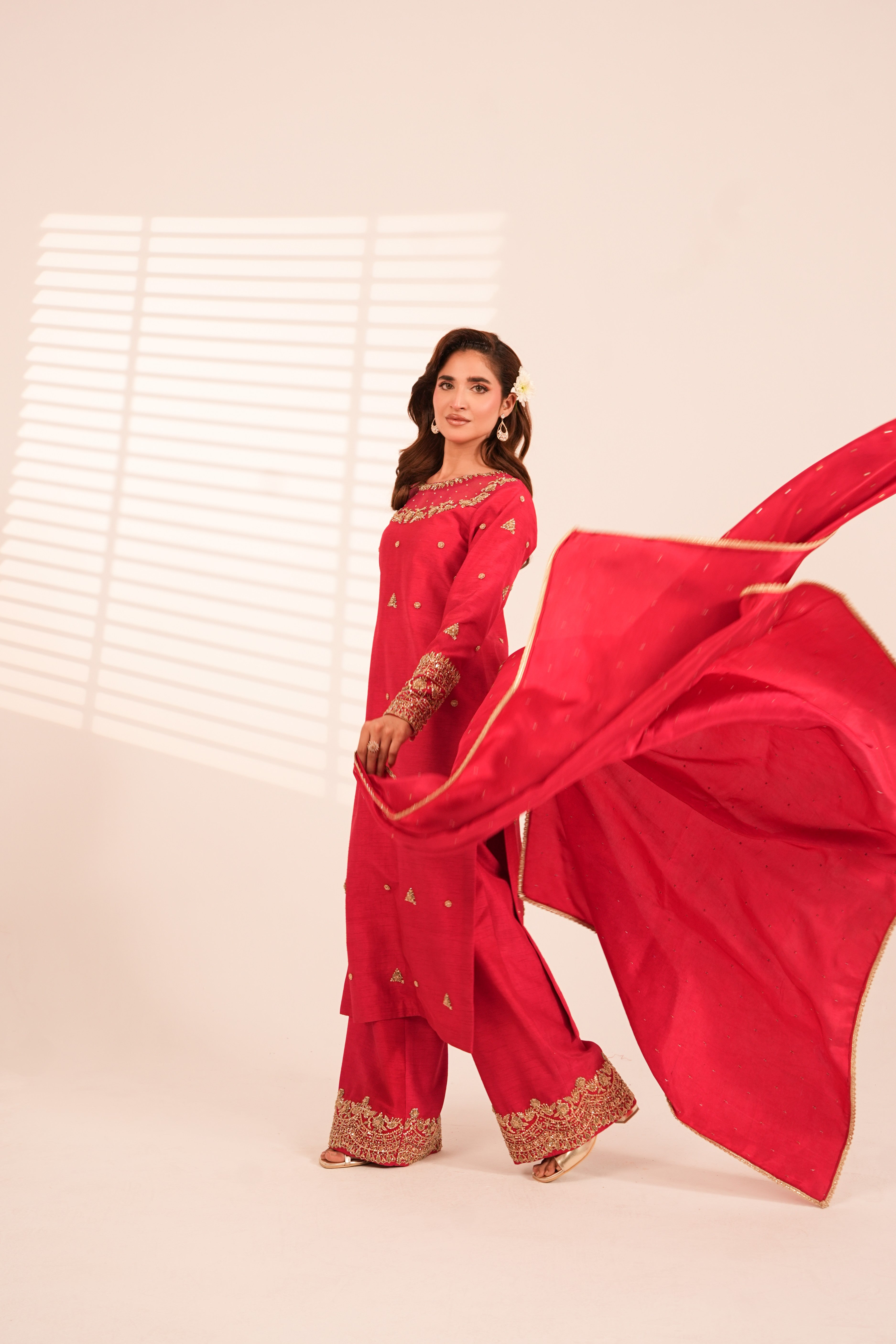 A woman modeling a bright red traditional outfit with gold embroidery and a matching flowing dupatta, posed against a warm off-white studio background with window blinds casting soft shadows.