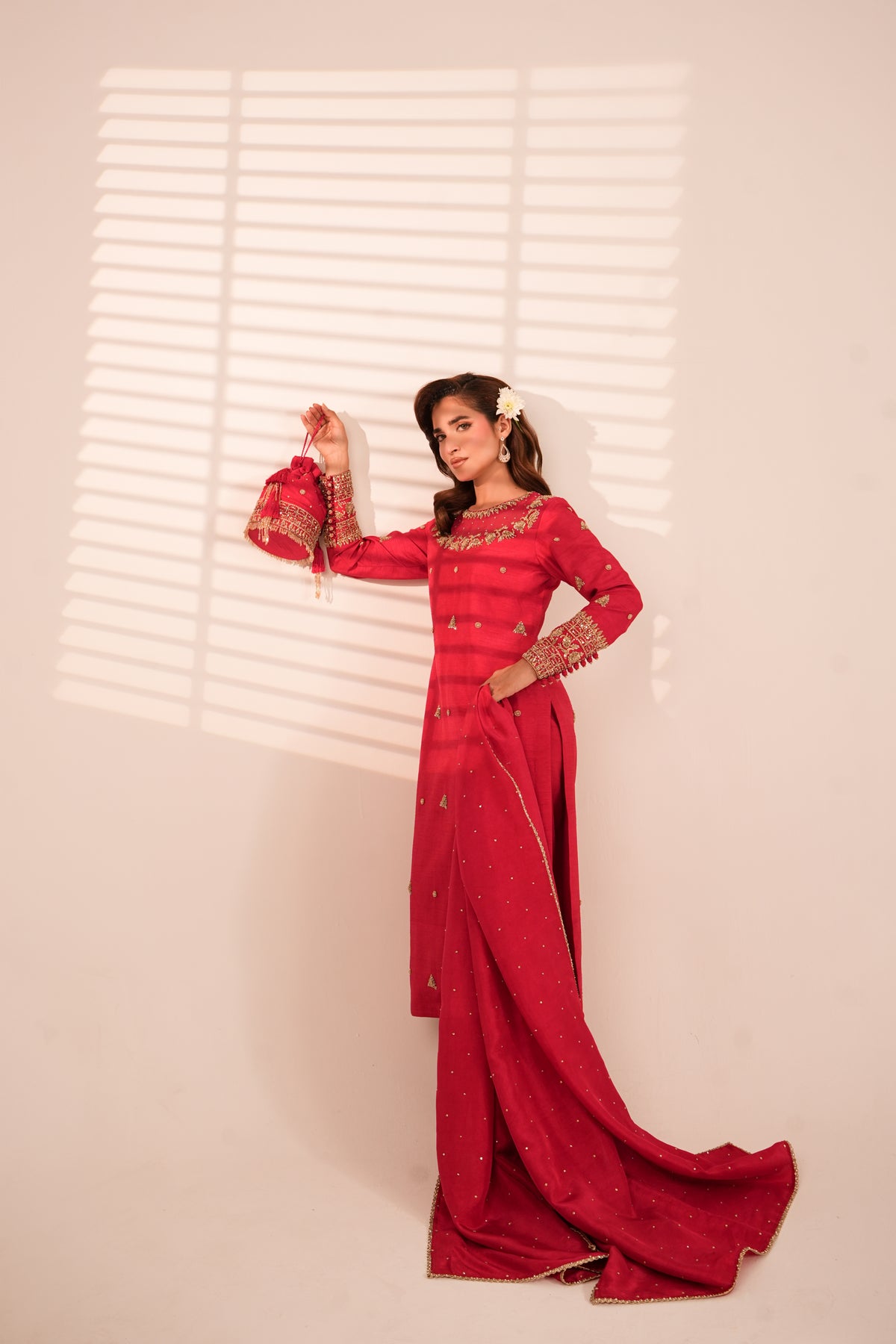 A woman models a long red traditional dress with gold embroidery and beadwork, standing against a light beige wall with window blind shadows.