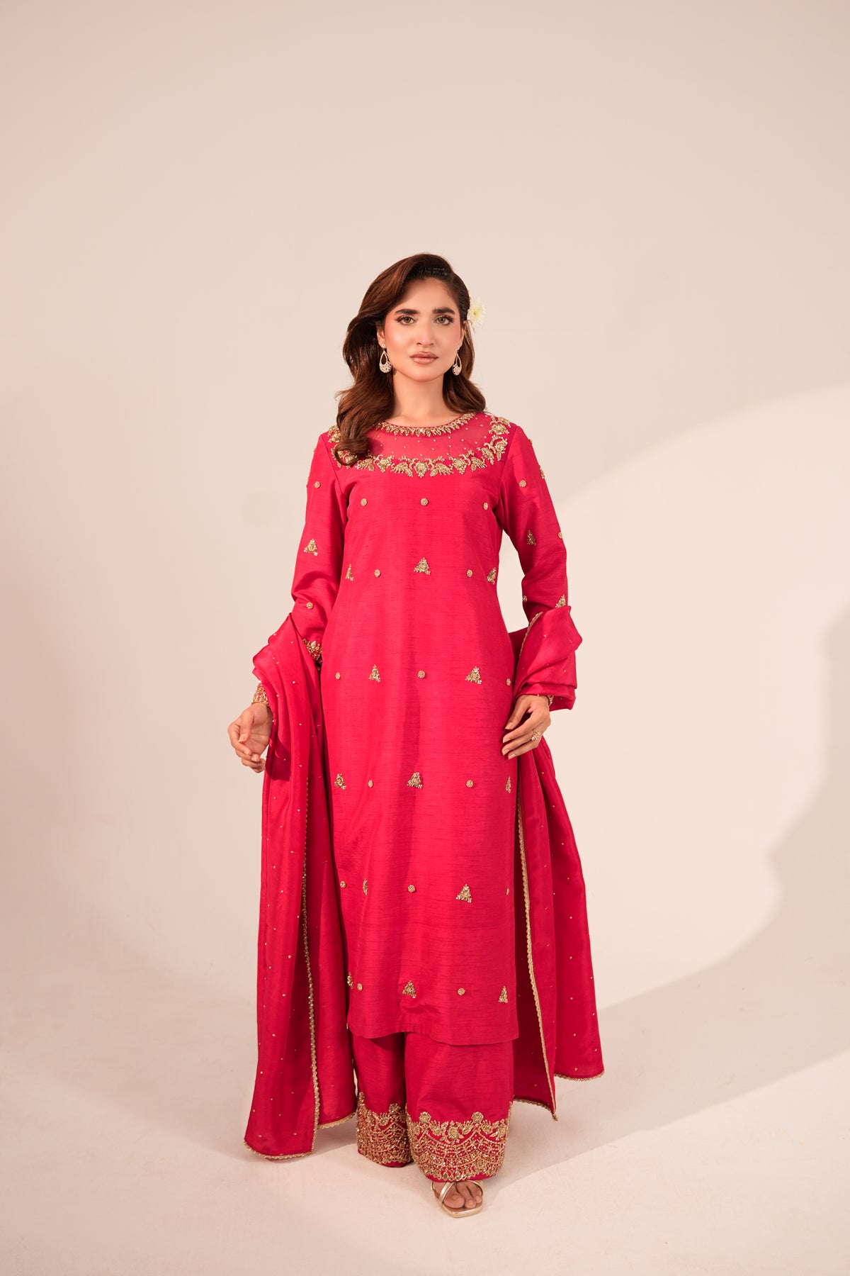 A woman modeling a bright pink traditional Indian outfit with gold embroidery, consisting of a long kurta, matching pants, and a draped dupatta, set against a plain beige studio background.