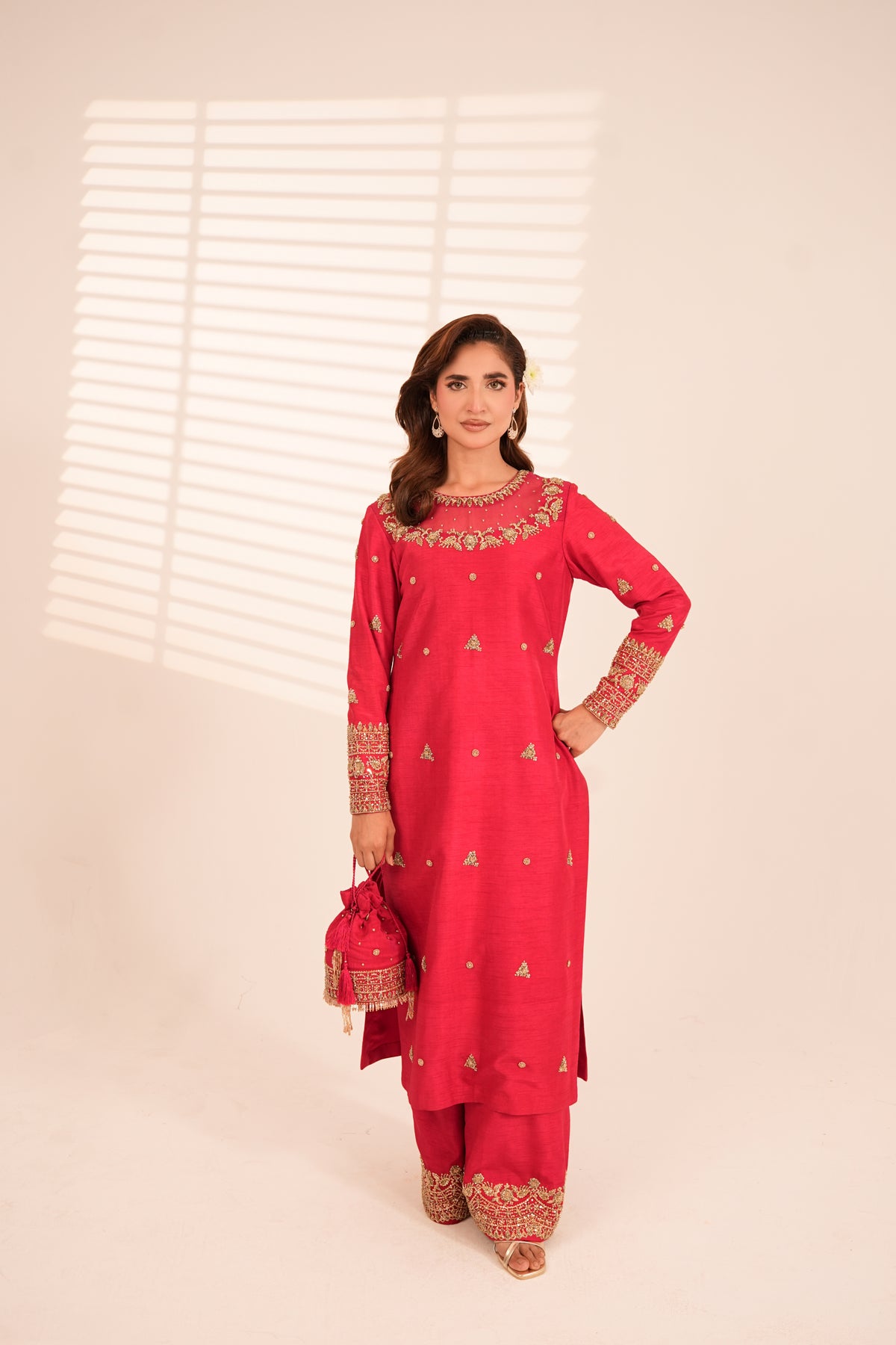 A woman modeling a bright pinkish-red traditional Indian outfit with gold embroidery, holding a matching embroidered drawstring pouch against a warm beige studio background with window blind shadows.