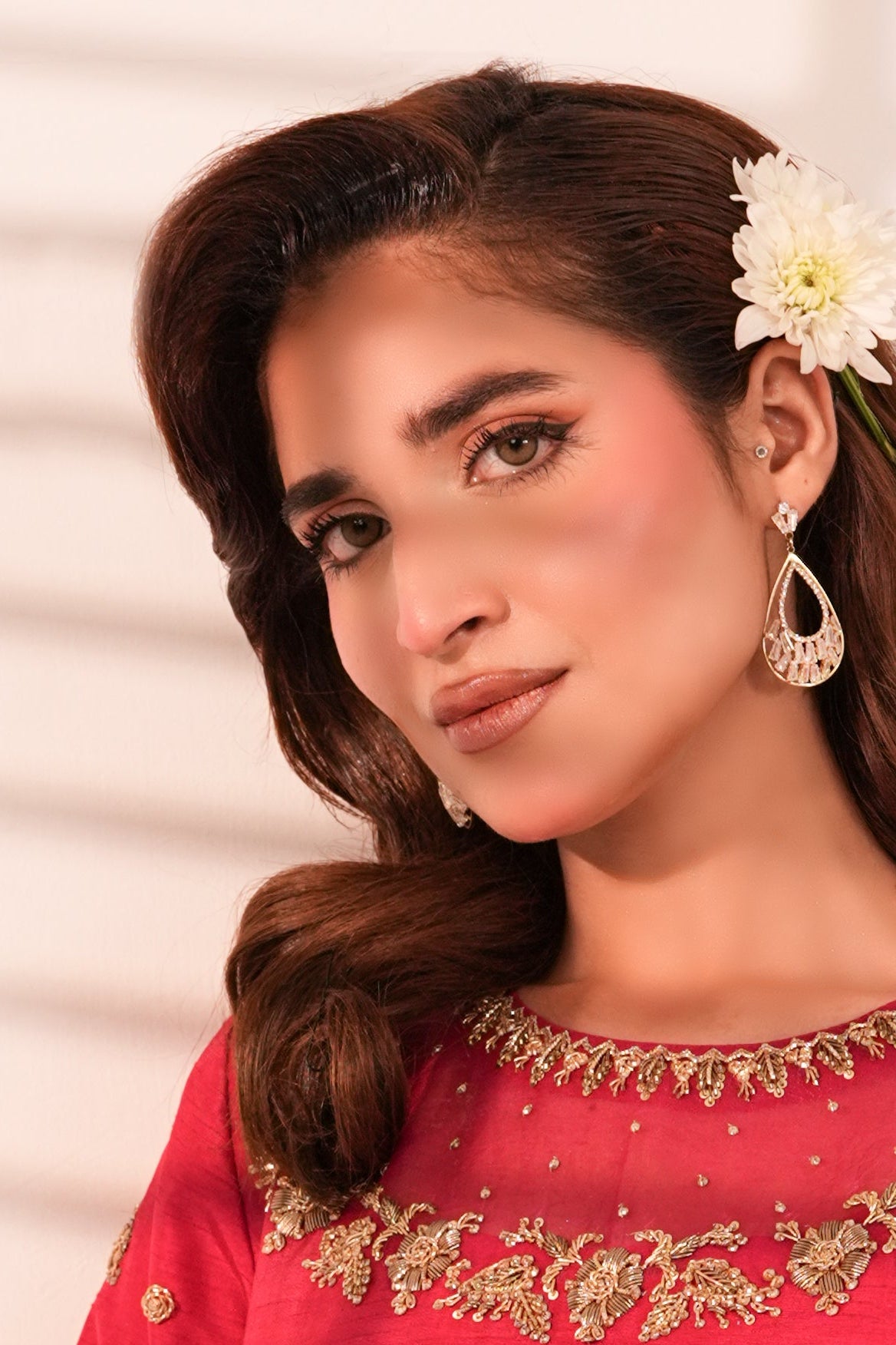 A woman with styled brown hair and a white flower accessory in her hair, wearing a red embroidered traditional outfit with gold detailing, set against a white wall with soft window shadow patterns.