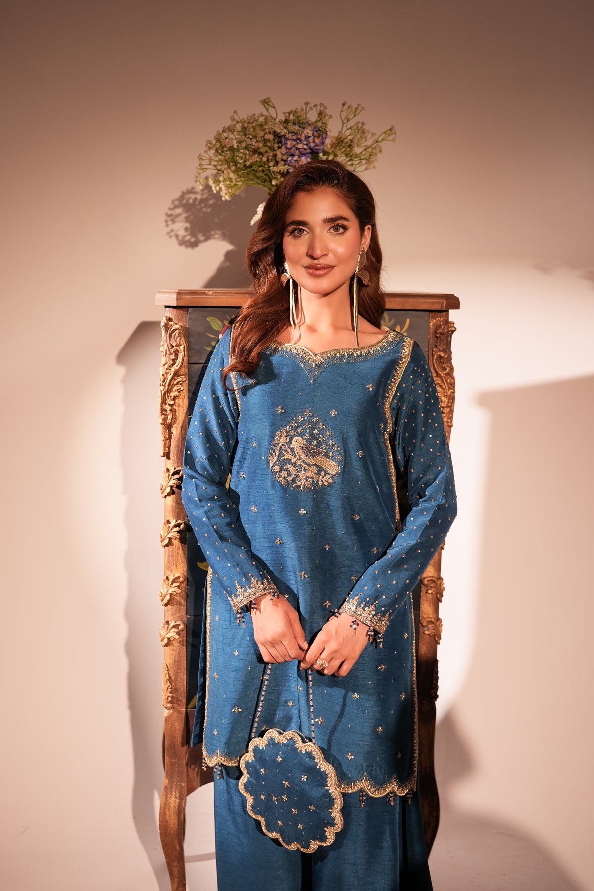 A woman models a blue traditional embroidered outfit with gold detailing, standing in front of a wooden chair against a beige wall.