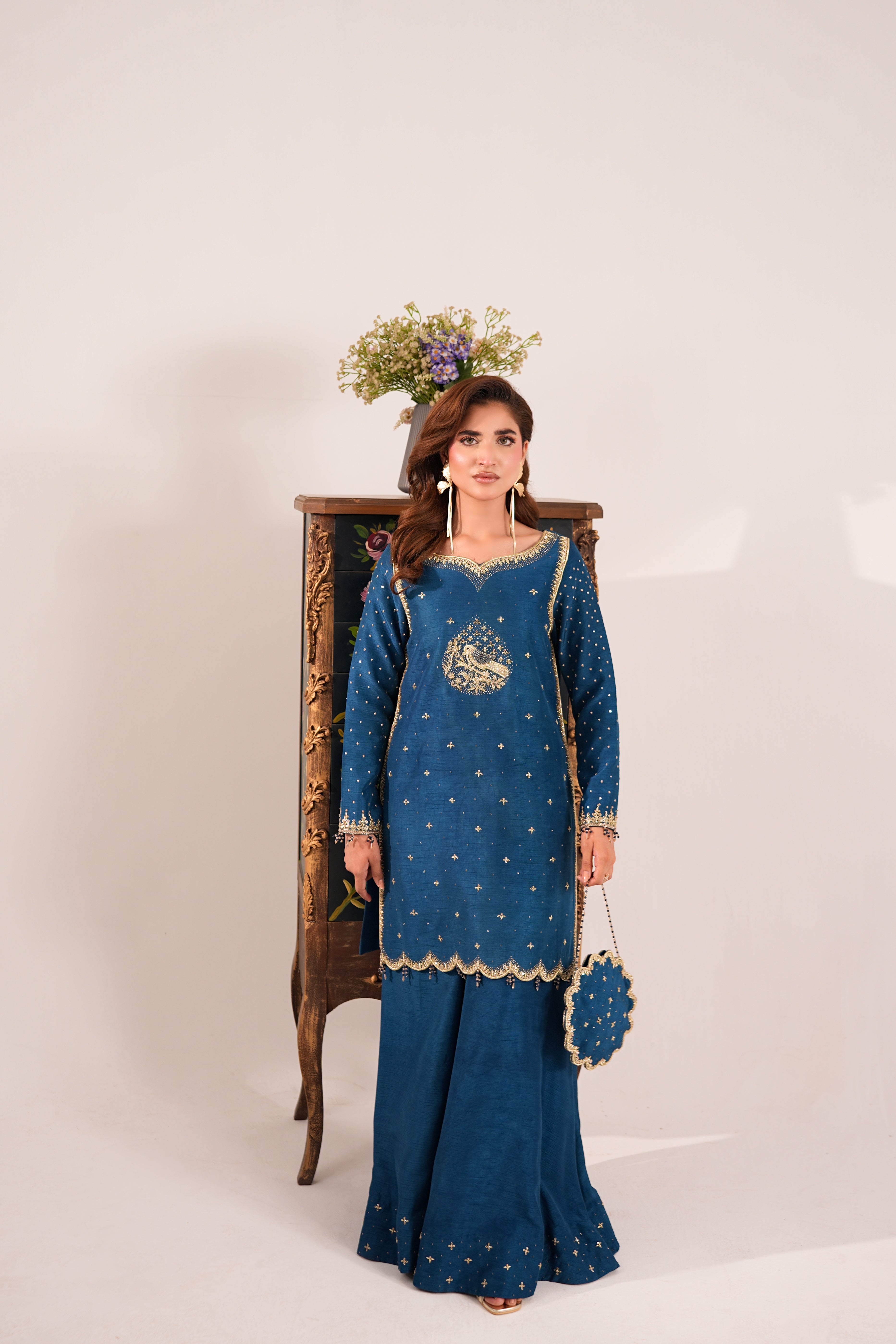 A woman models a traditional blue ethnic outfit with gold embroidery, standing in front of a vintage wooden cabinet against a plain light background.