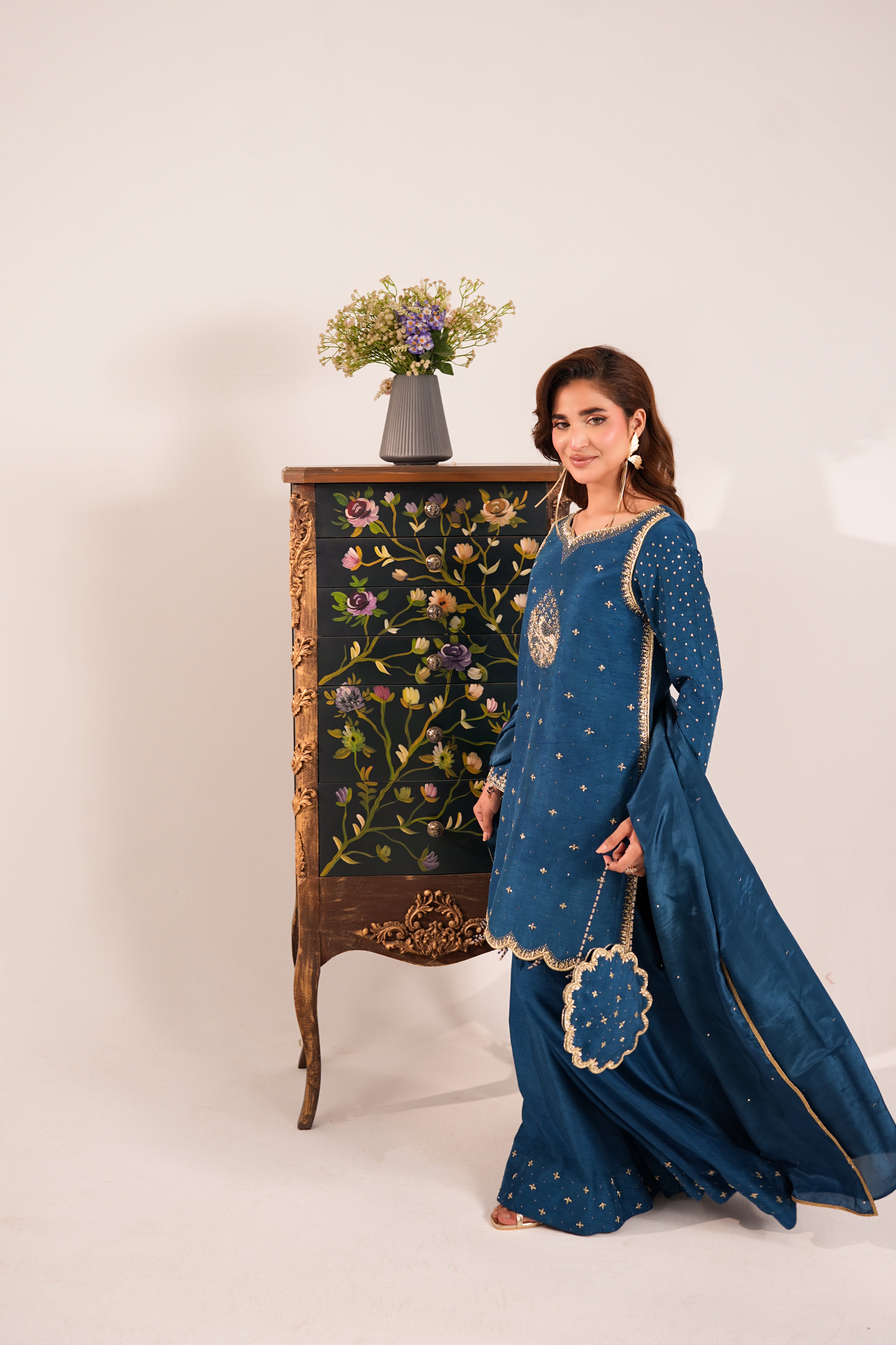 A woman models a traditional blue embroidered salwar kameez set with a matching dupatta, standing next to an ornate floral cabinet against a plain studio background.