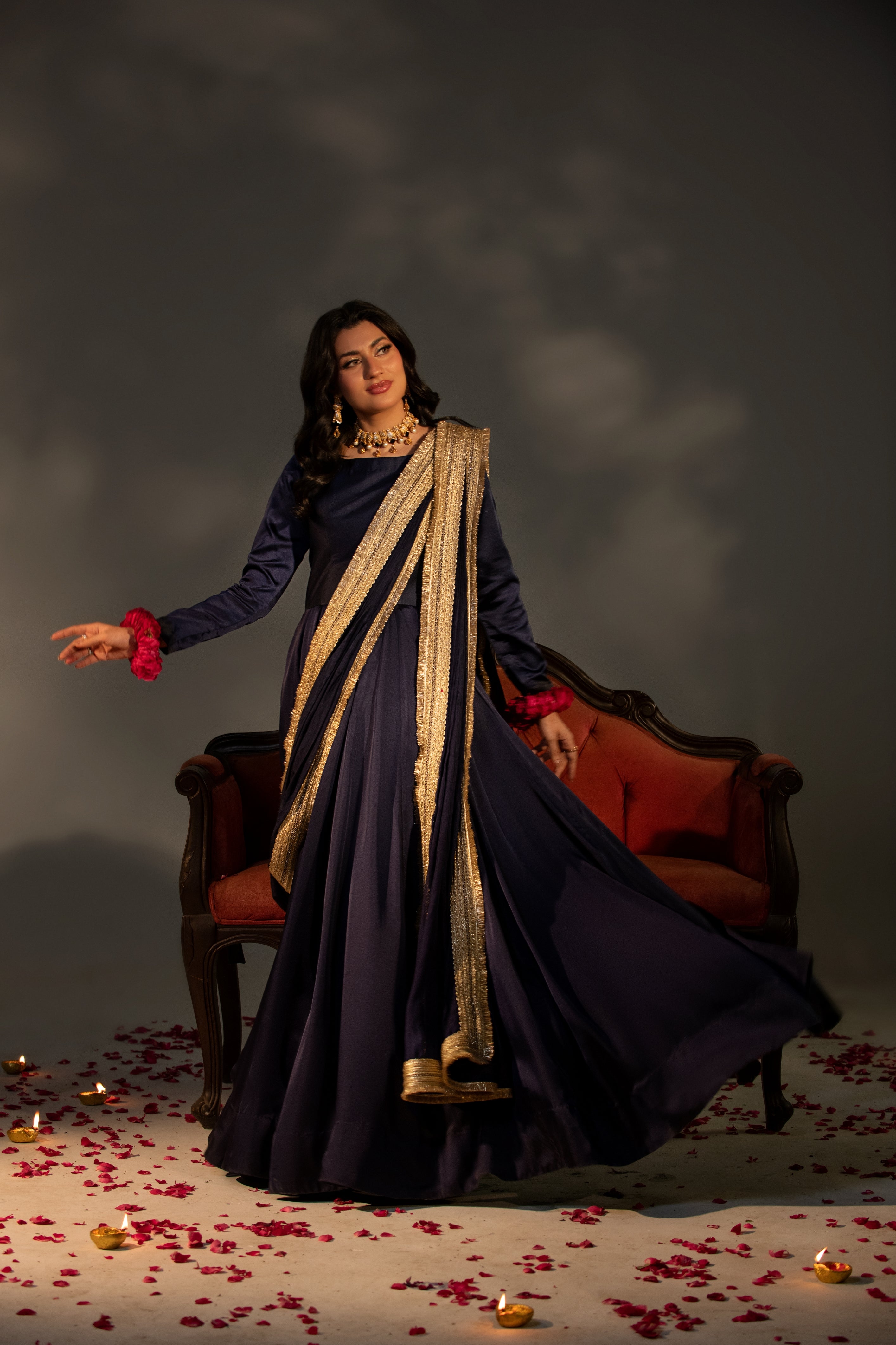 Woman in a traditional outfit standing in a room with red chairs and scattered rose petals on the floor.