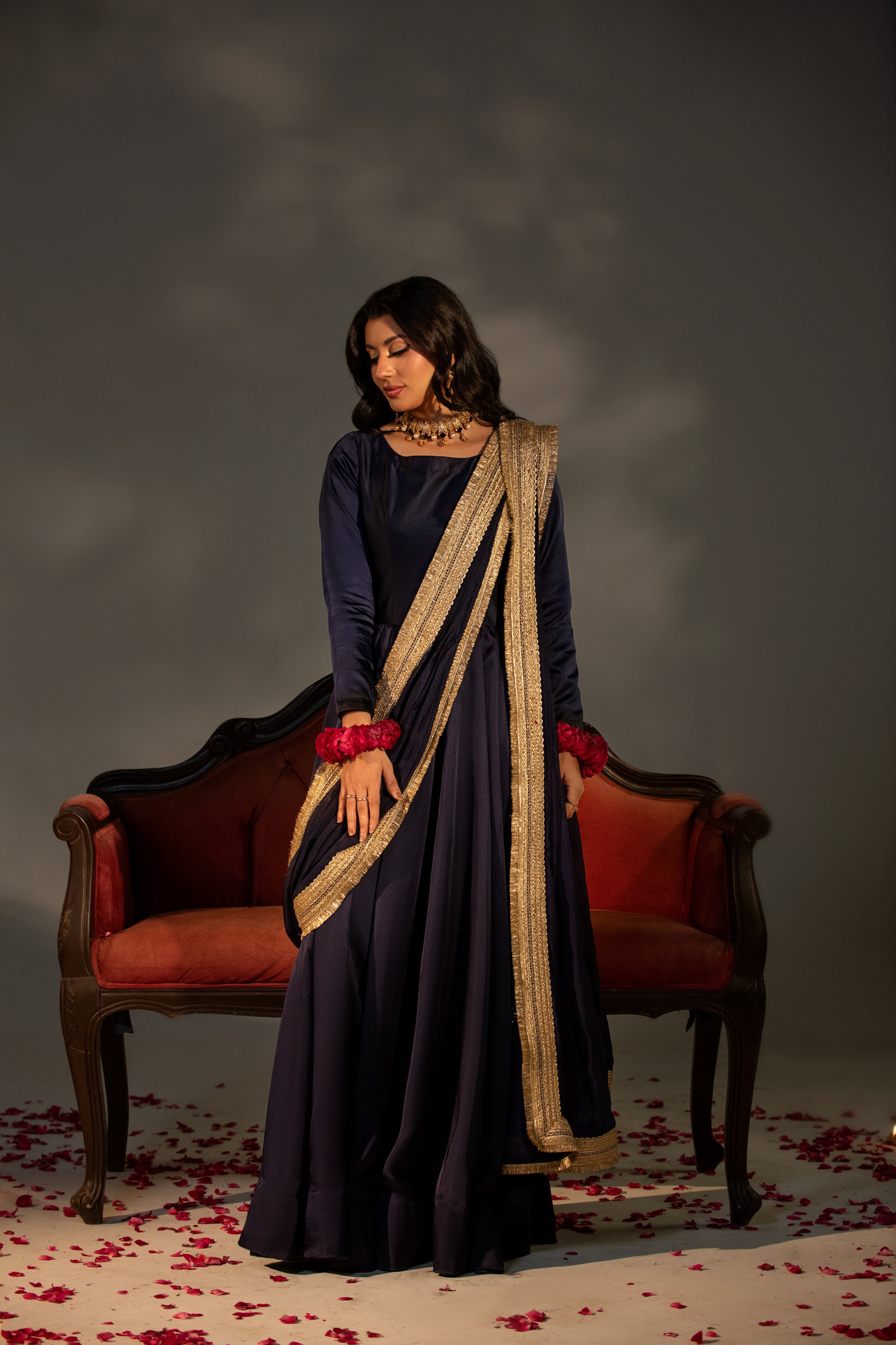 Woman in a dark blue traditional outfit with gold border standing in front of a red velvet couch.