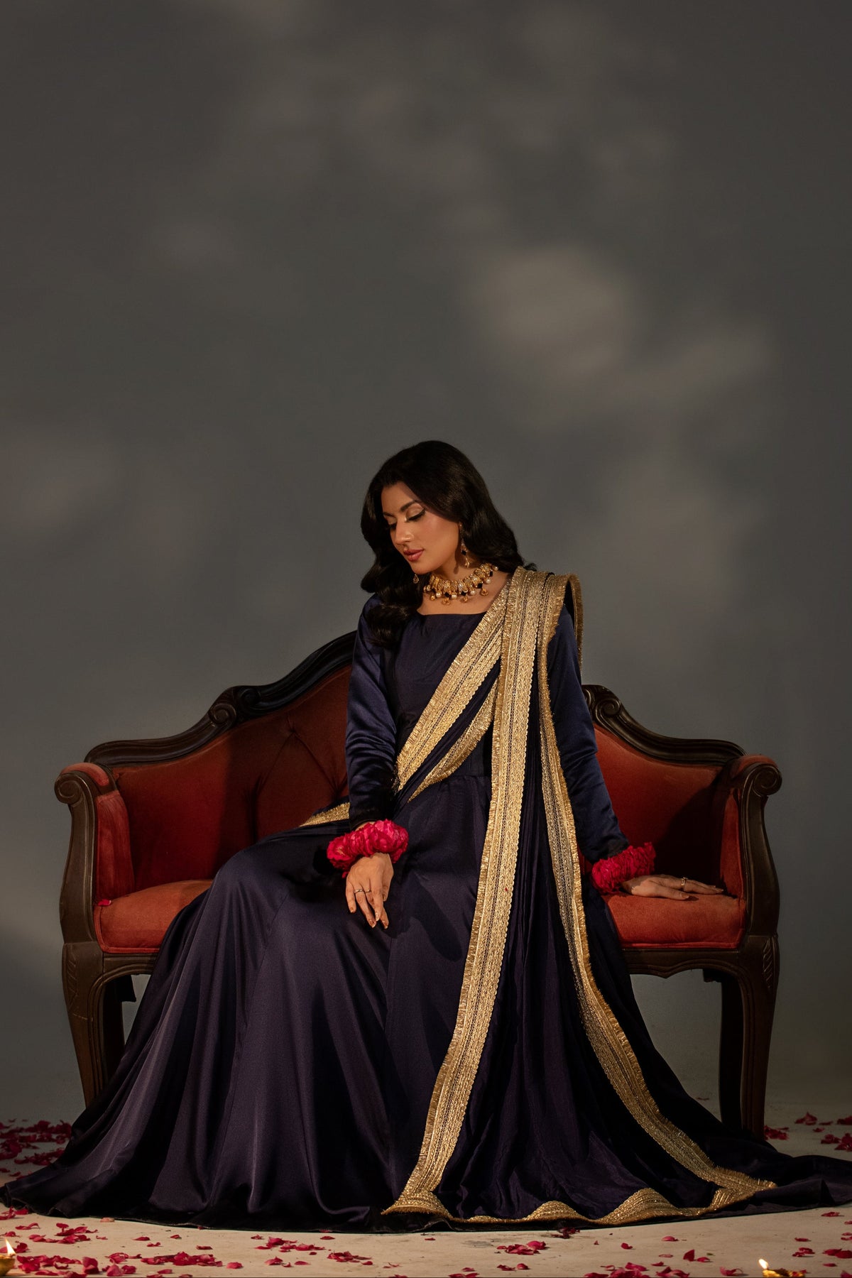 Woman in a dark blue dress with gold accents sitting on a red couch against a dark background