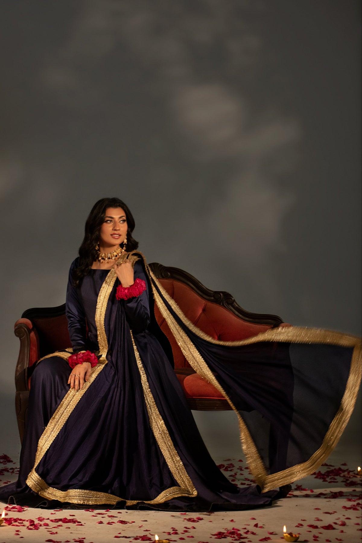 Woman in a dark blue saree with gold border sitting on a red couch against a dark background
