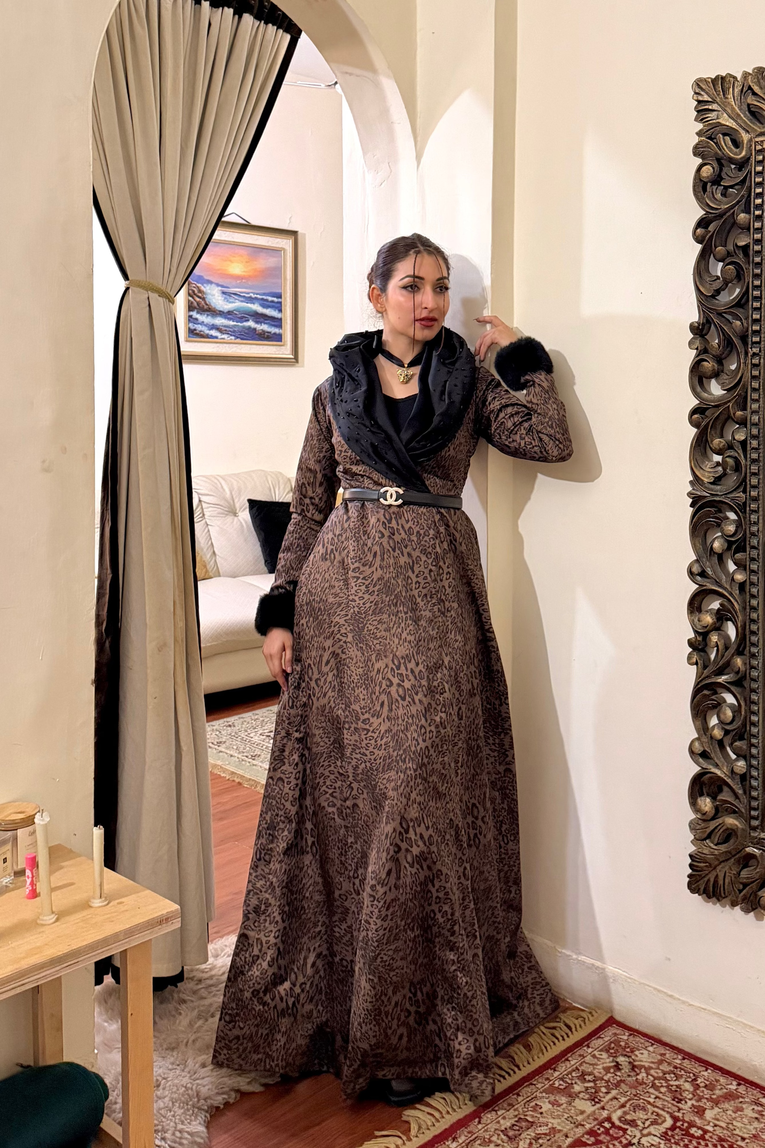 Woman in a long brown dress standing in a room with a large mirror and decorative elements.