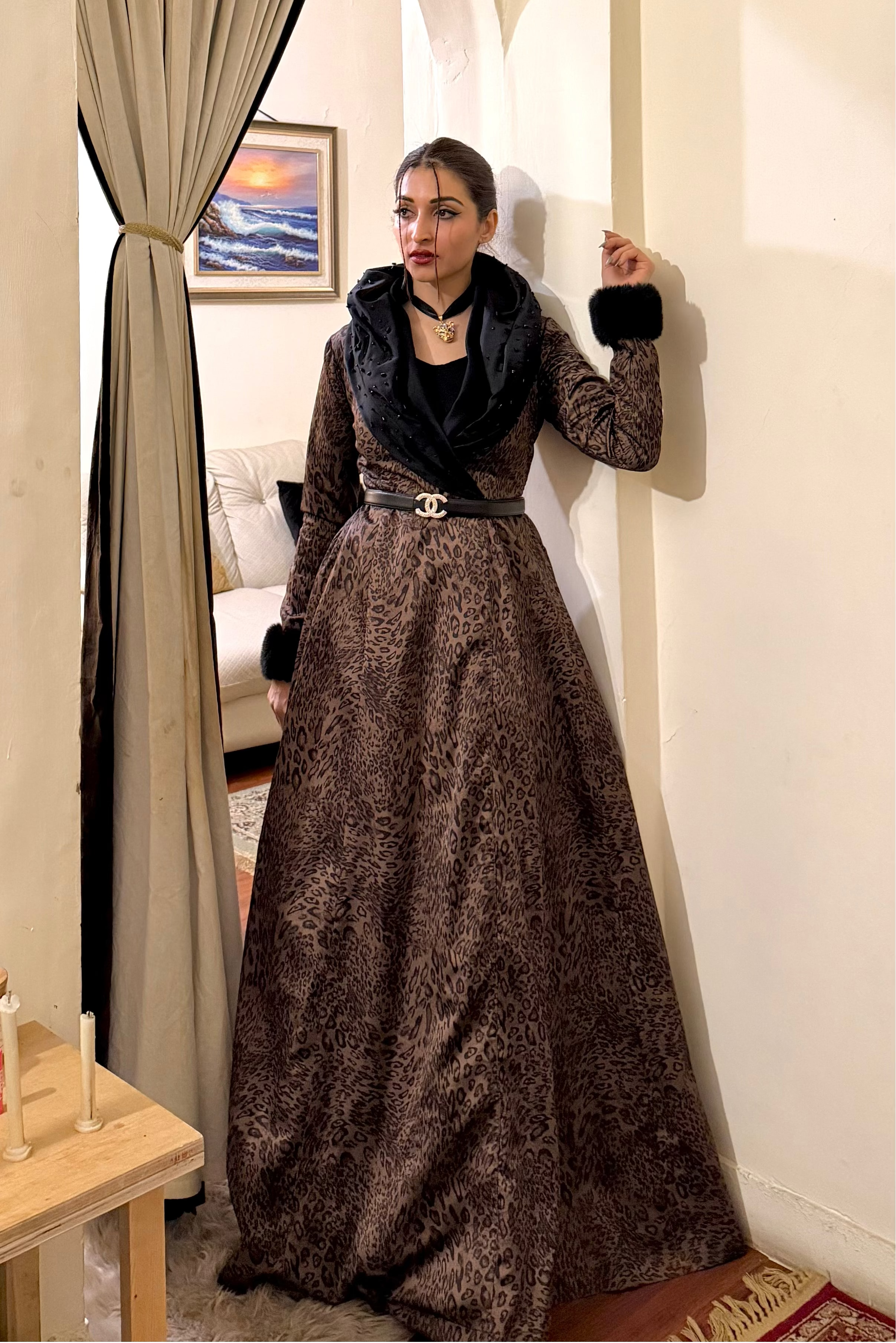 Woman in a long, dark brown dress standing in front of a decorative mirror in a room.