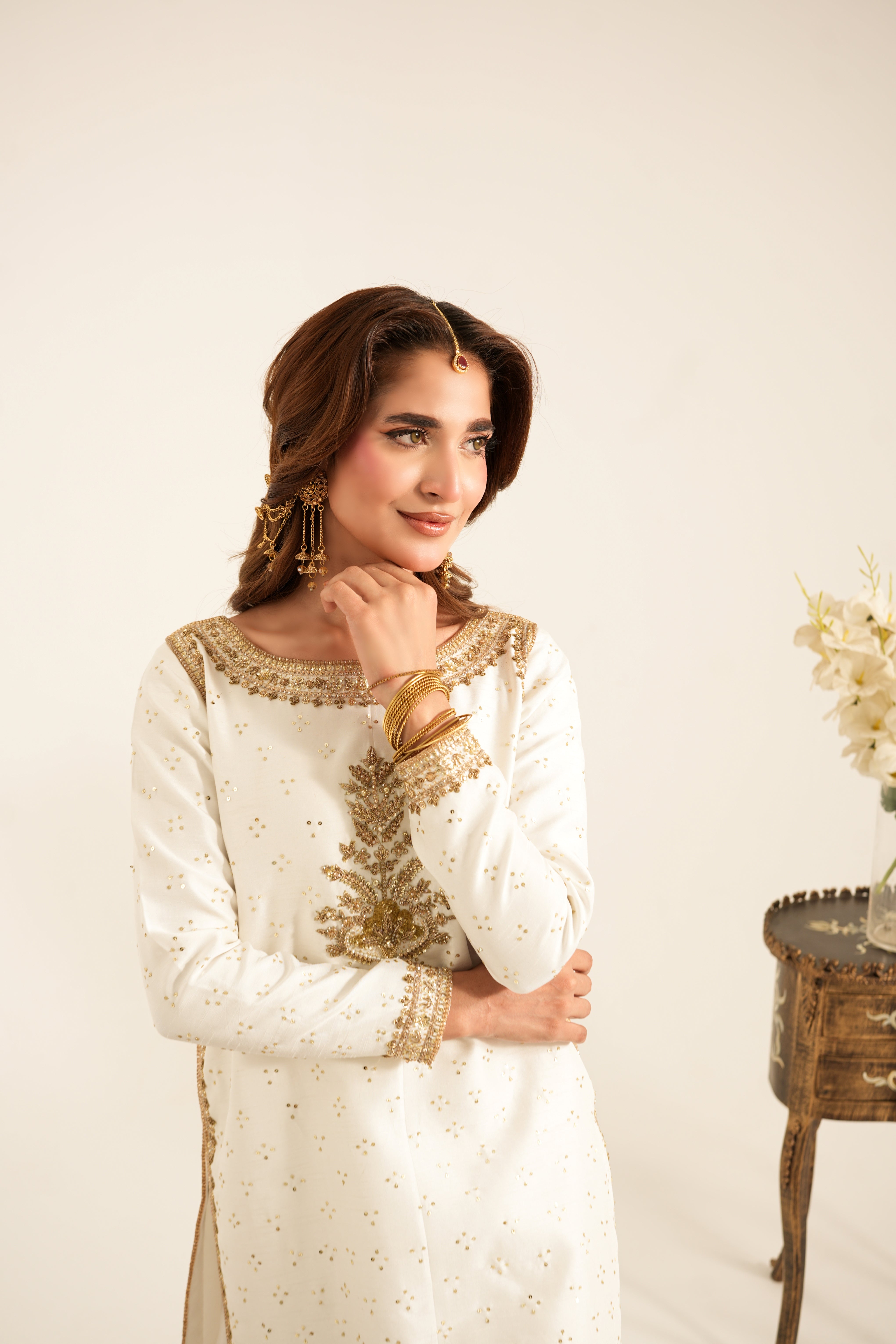 A woman wearing a white traditional embroidered kurta with gold detailing, styled with gold jewelry, standing against a warm off-white studio background next to a small wooden table with flowers.