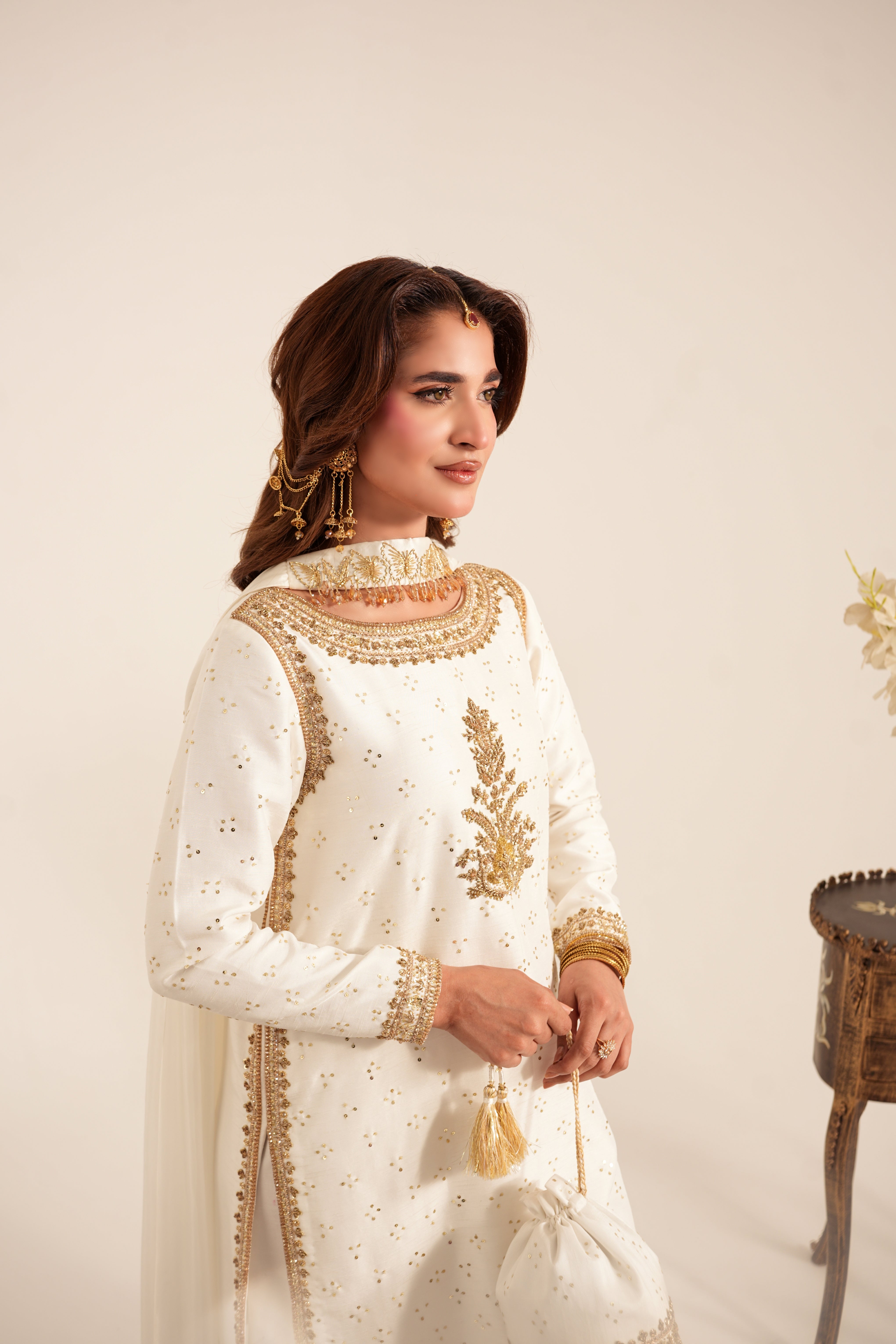 A woman wearing a traditional white embroidered kurta with gold detailing, holding a matching drawstring bag against a warm beige studio background.