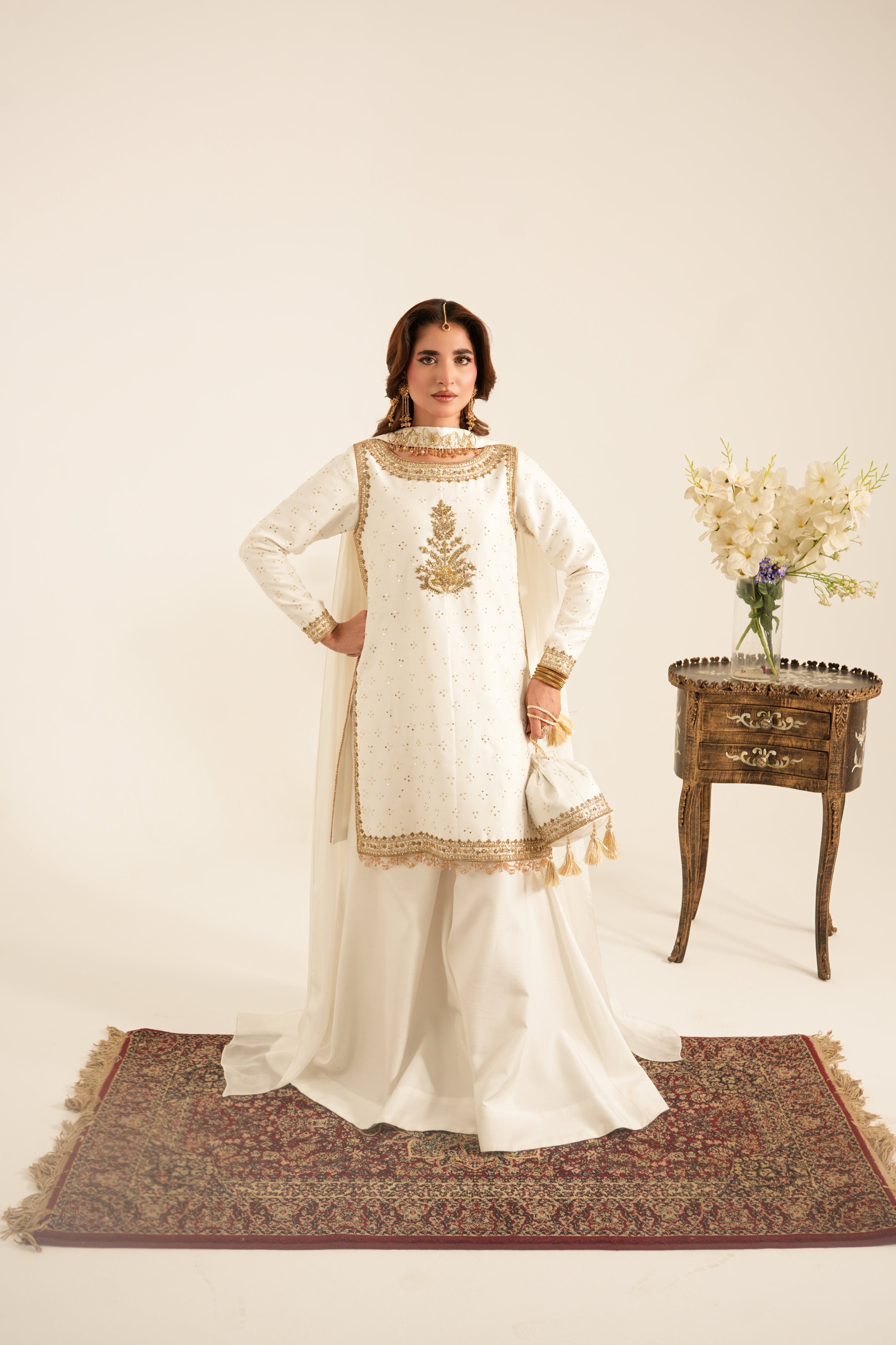 A woman models a traditional white three-piece ethnic suit with gold embroidery, standing on a patterned rug next to a small wooden side table with flowers, against a solid cream background.