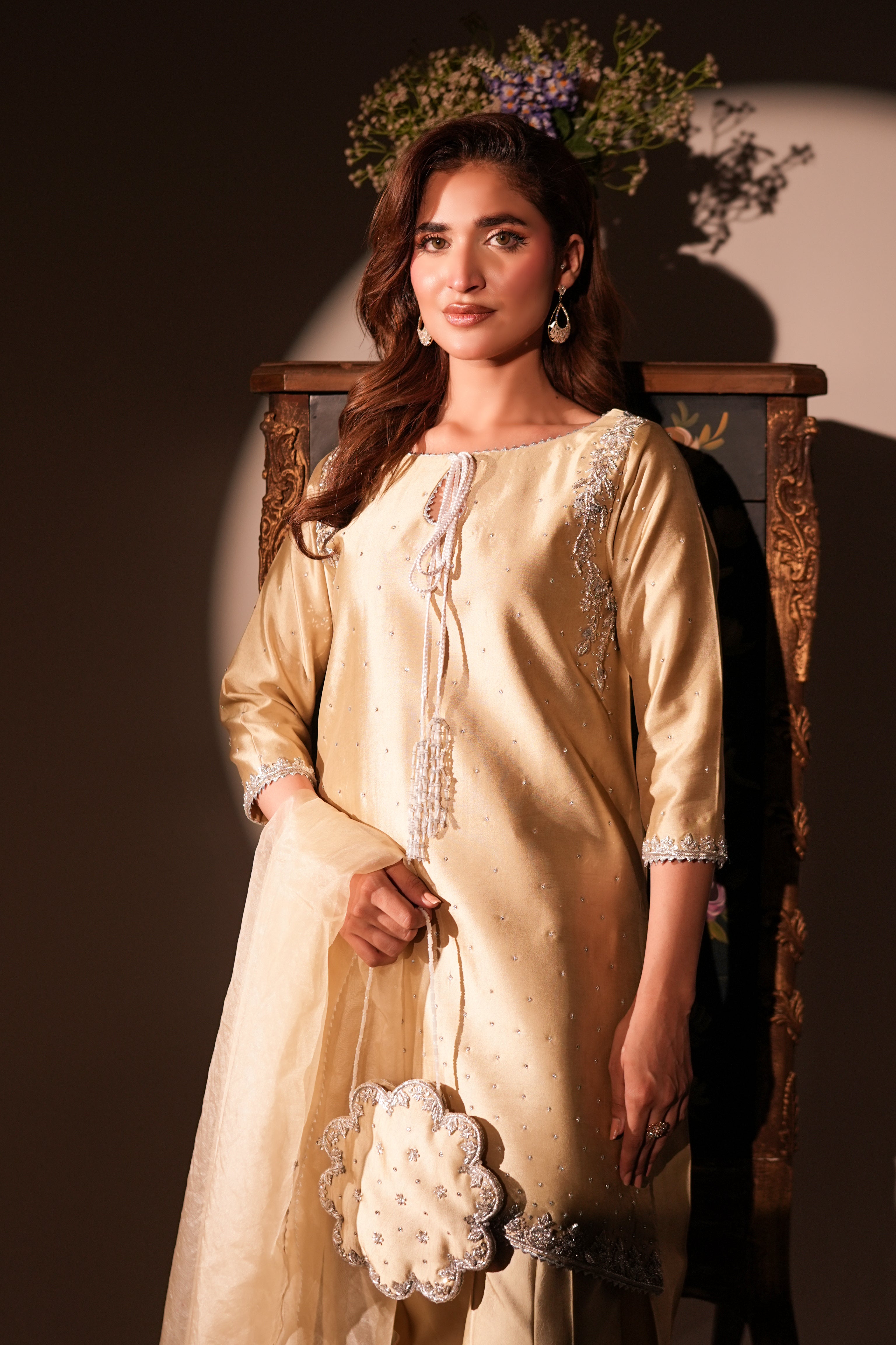A woman models a traditional beige embroidered kurta set with a matching dupatta, standing in front of a dark wooden chair under dramatic warm lighting.