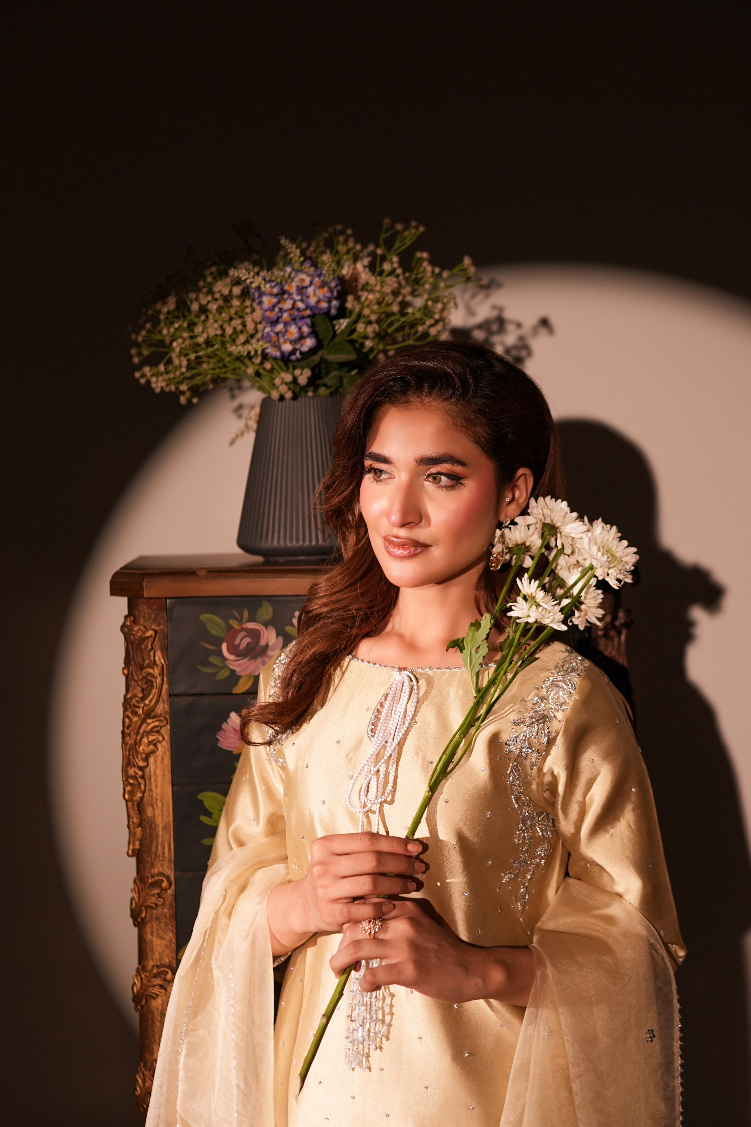 A woman wearing a cream-colored embroidered traditional outfit holds white flowers, standing in front of a dark background with a spotlight effect and a decorative cabinet.