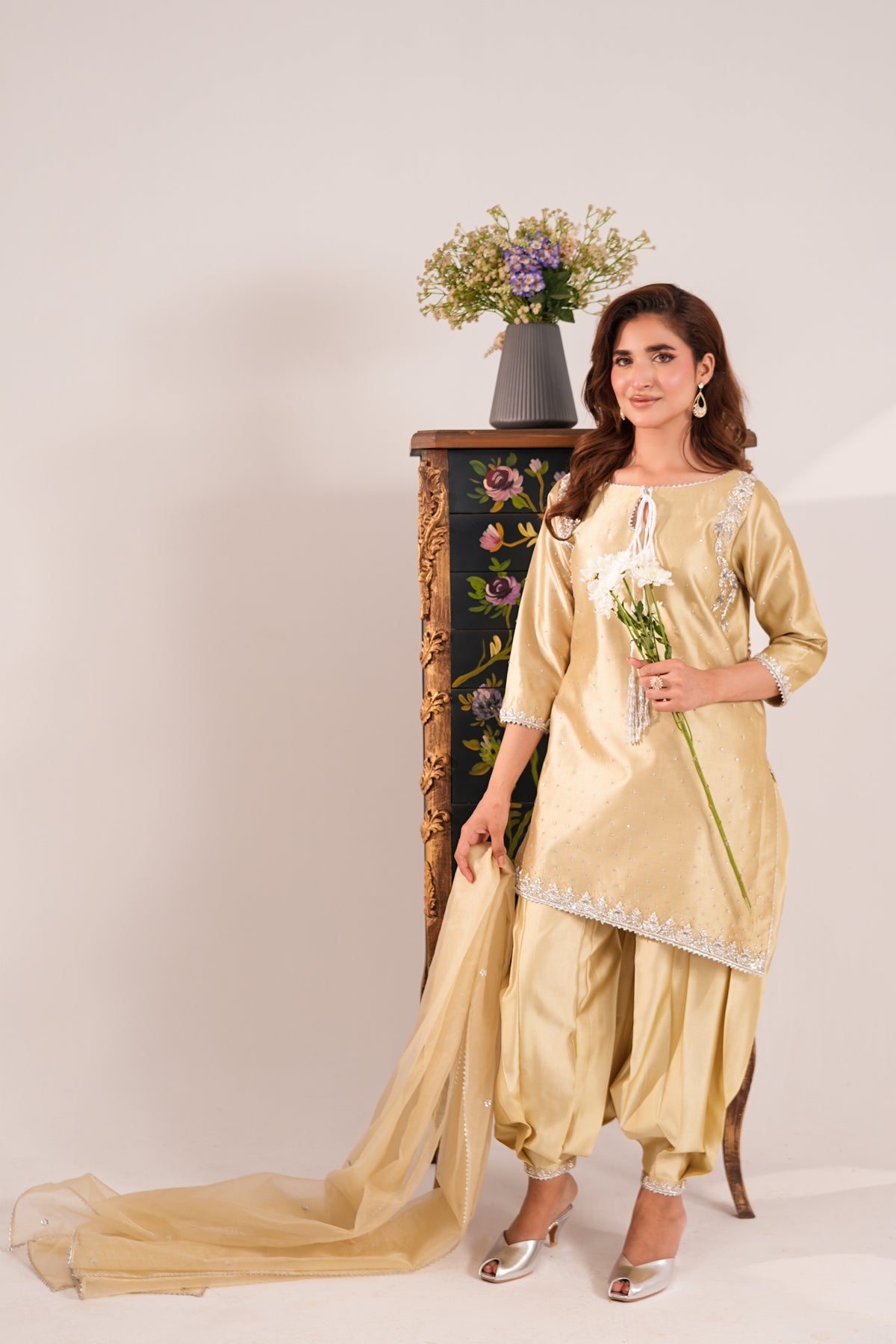 A woman models a traditional South Asian salwar kameez suit in a light gold/champagne color with white embroidery. The set includes a long kurta, matching pants, and a sheer dupatta draped on the floor.