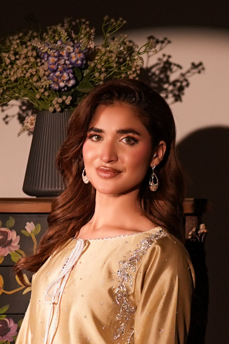 A woman wearing a cream-colored embroidered kurta stands in front of a spotlighted dark background, with a floral arrangement and a cabinet visible behind her.