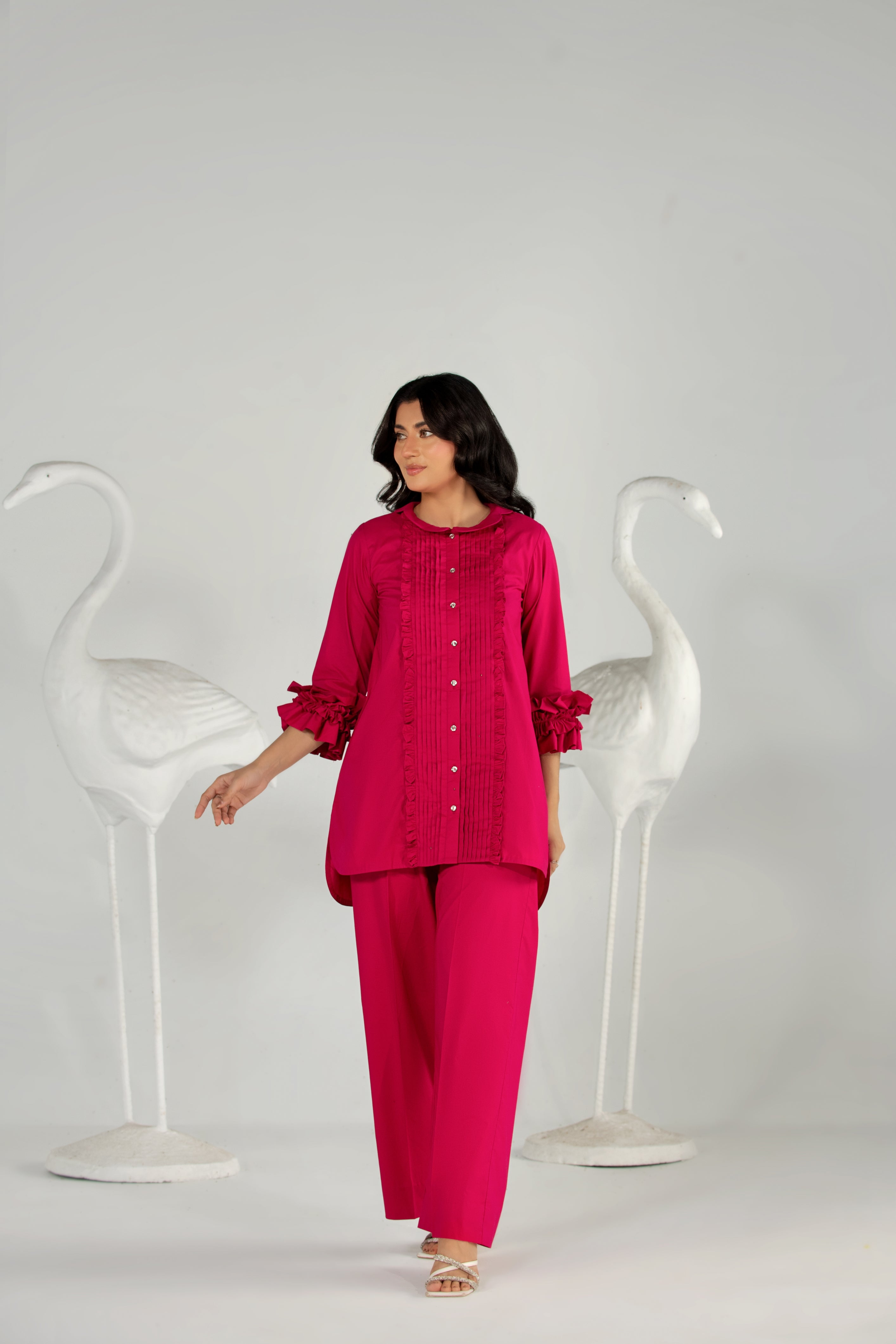 Woman wearing a bright pink outfit standing between two white chairs on a plain background