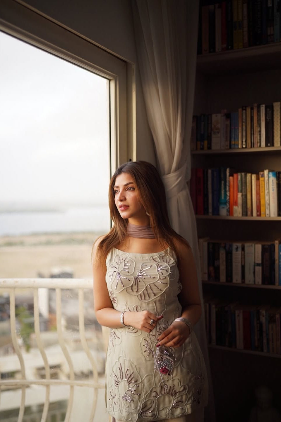 Woman in a floral dress standing by a window with a view of a field, next to a bookshelf.