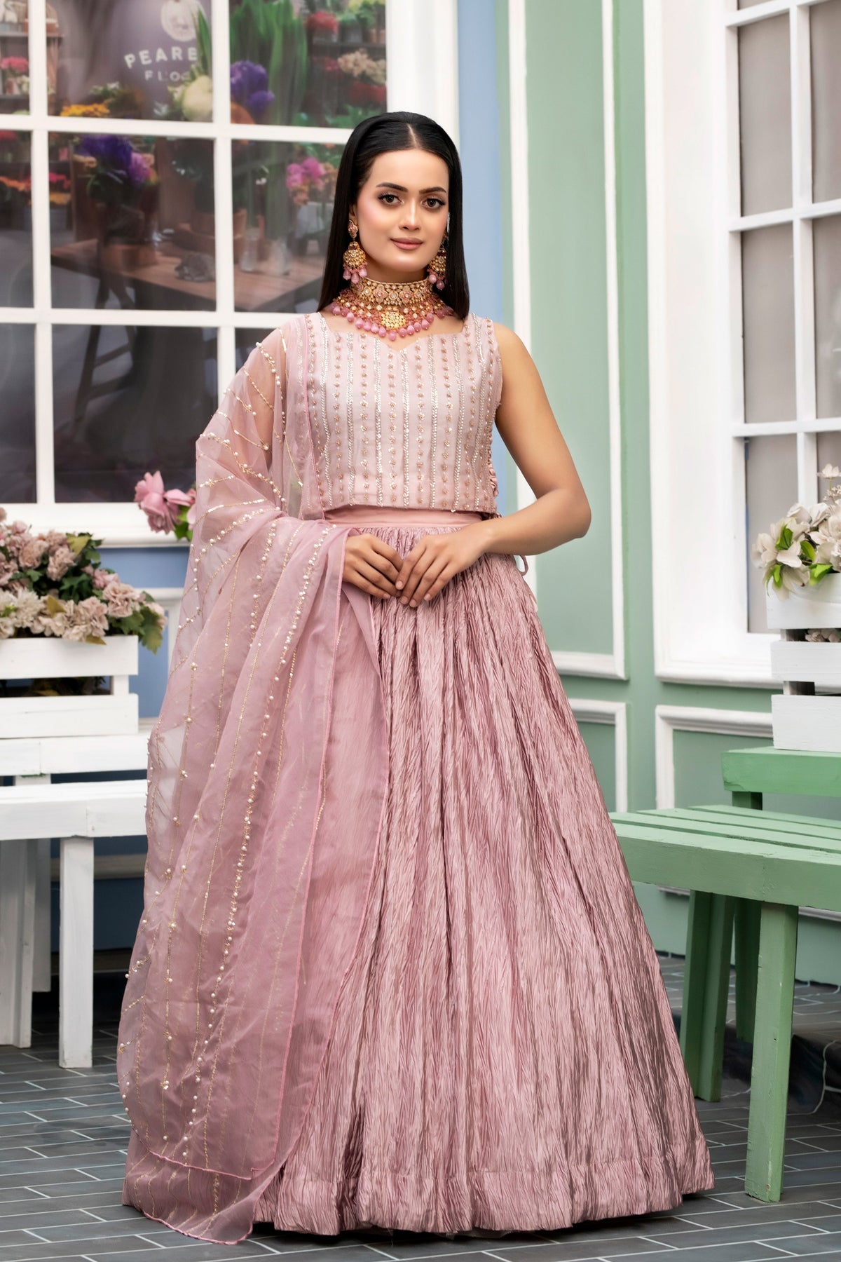 Woman in a pink traditional outfit standing in an outdoor setting with flowers and furniture.