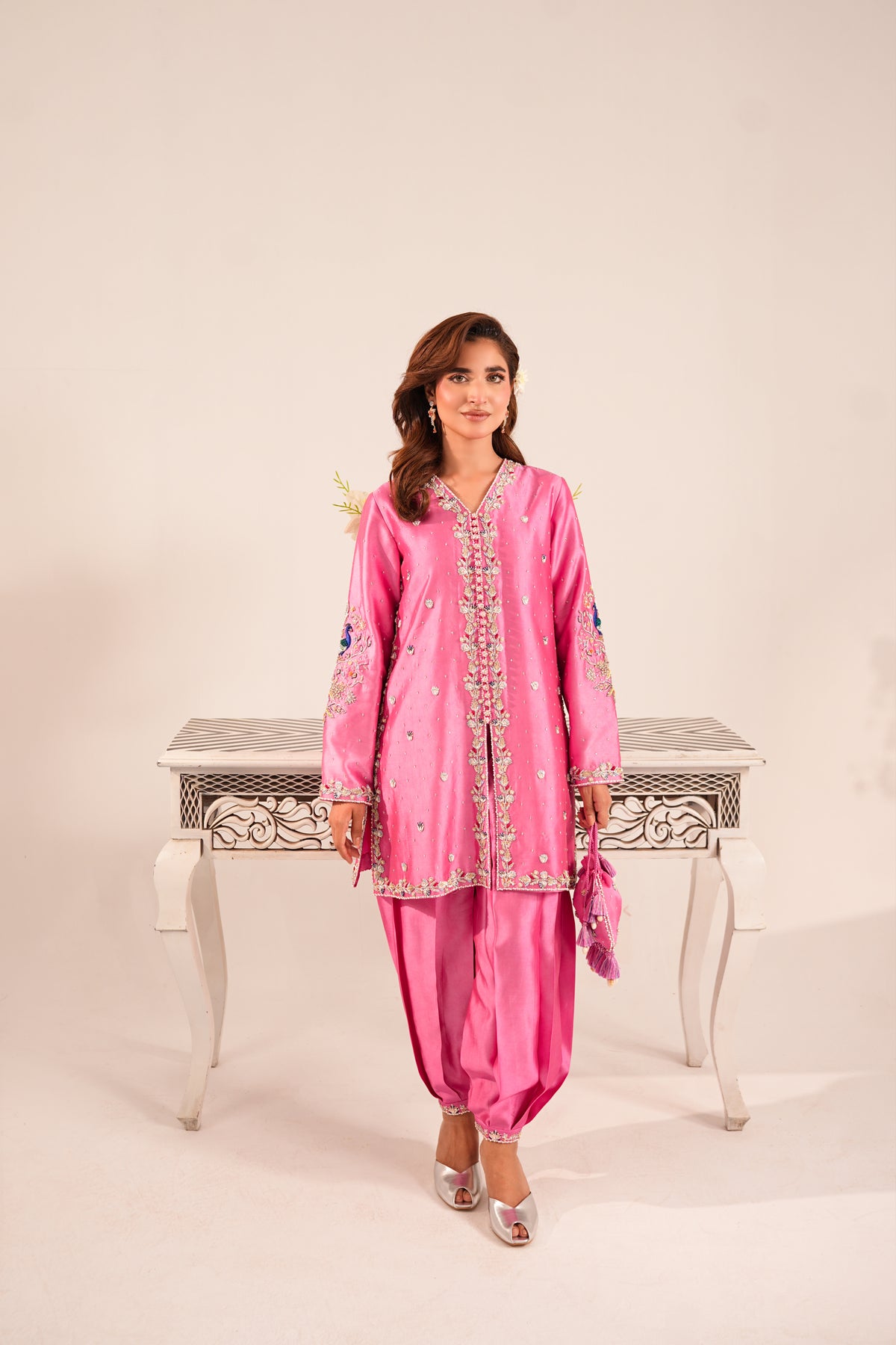 A woman models a bright pink traditional South Asian outfit with matching pants and a matching pink dupatta, standing in front of a white decorative table against a light beige studio background.