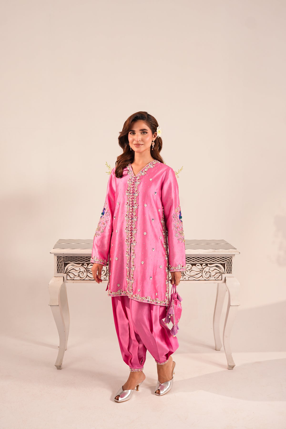 A woman modeling a pink traditional South Asian outfit with embroidered details, standing in front of a light beige studio backdrop and a white decorative table.