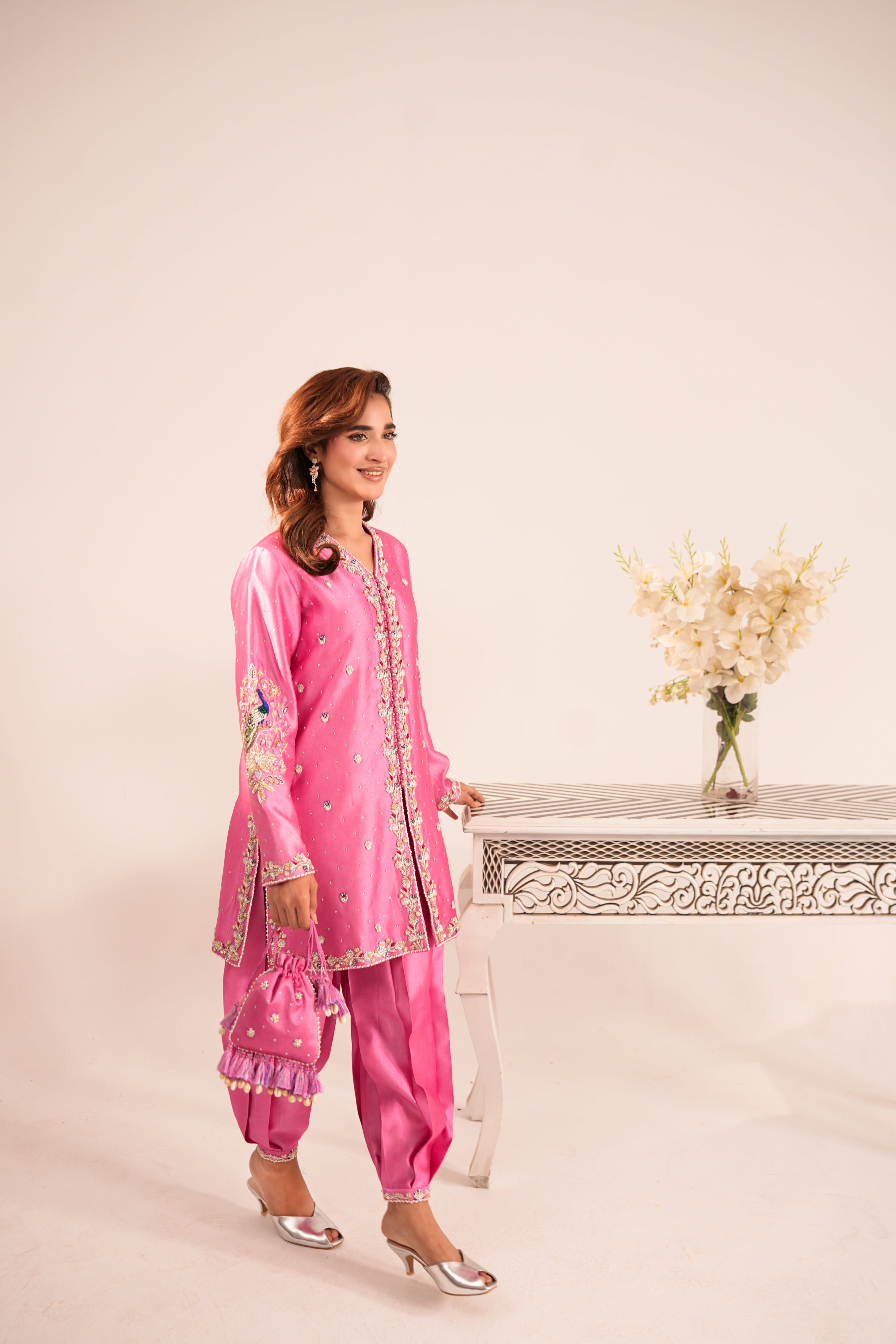 A woman models a bright pink traditional South Asian outfit with matching pants, a long embroidered kurta, and metallic silver heels, standing next to a decorative table with flowers against a plain beige background.
