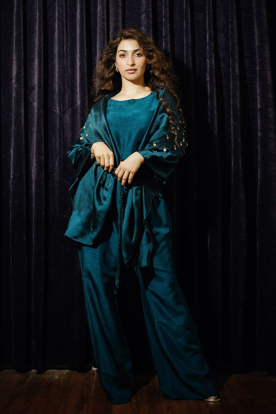 Woman in a teal outfit standing against a dark curtain