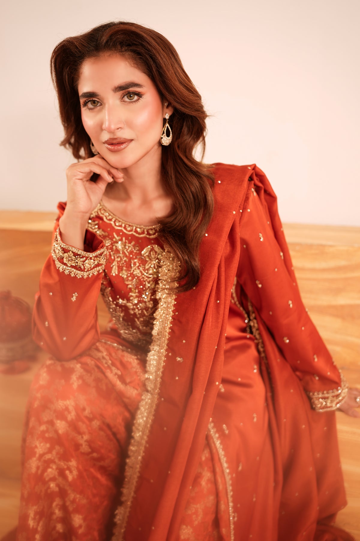 A woman wearing a traditional South Asian outfit in rust orange and gold, posing indoors against a neutral beige background.