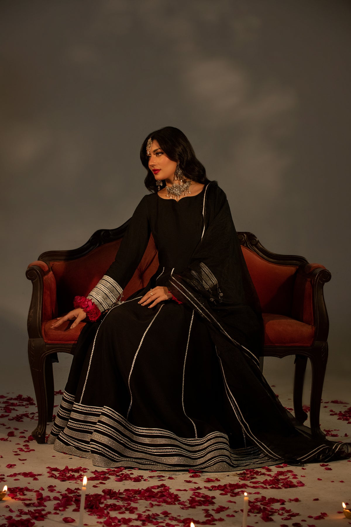 Woman in a black dress sitting on a red couch with candles and rose petals on the floor.