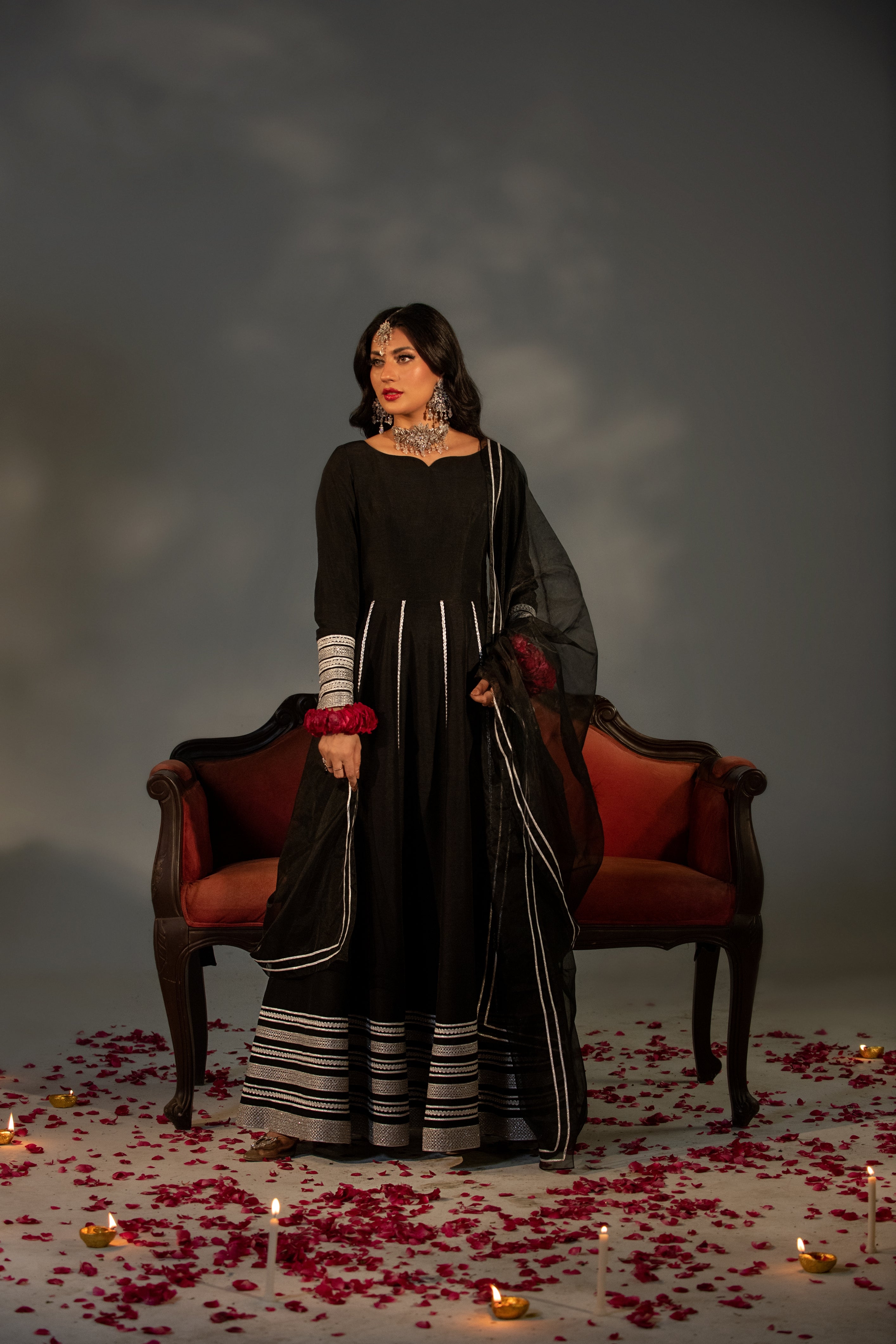Woman in a black traditional outfit standing in front of a red chair with candles and rose petals on the floor.