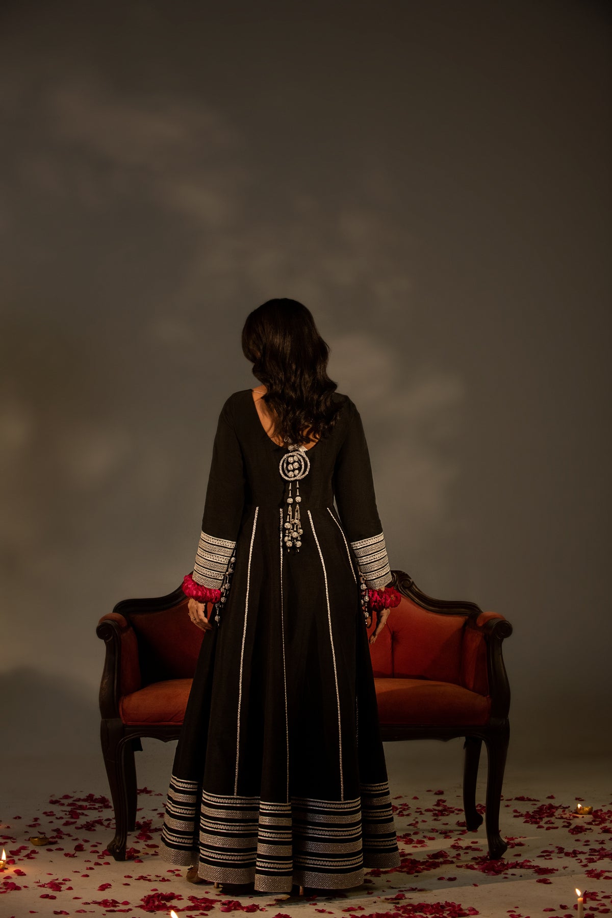 Woman in a long black dress standing in front of a red chair with a dark, moody background.