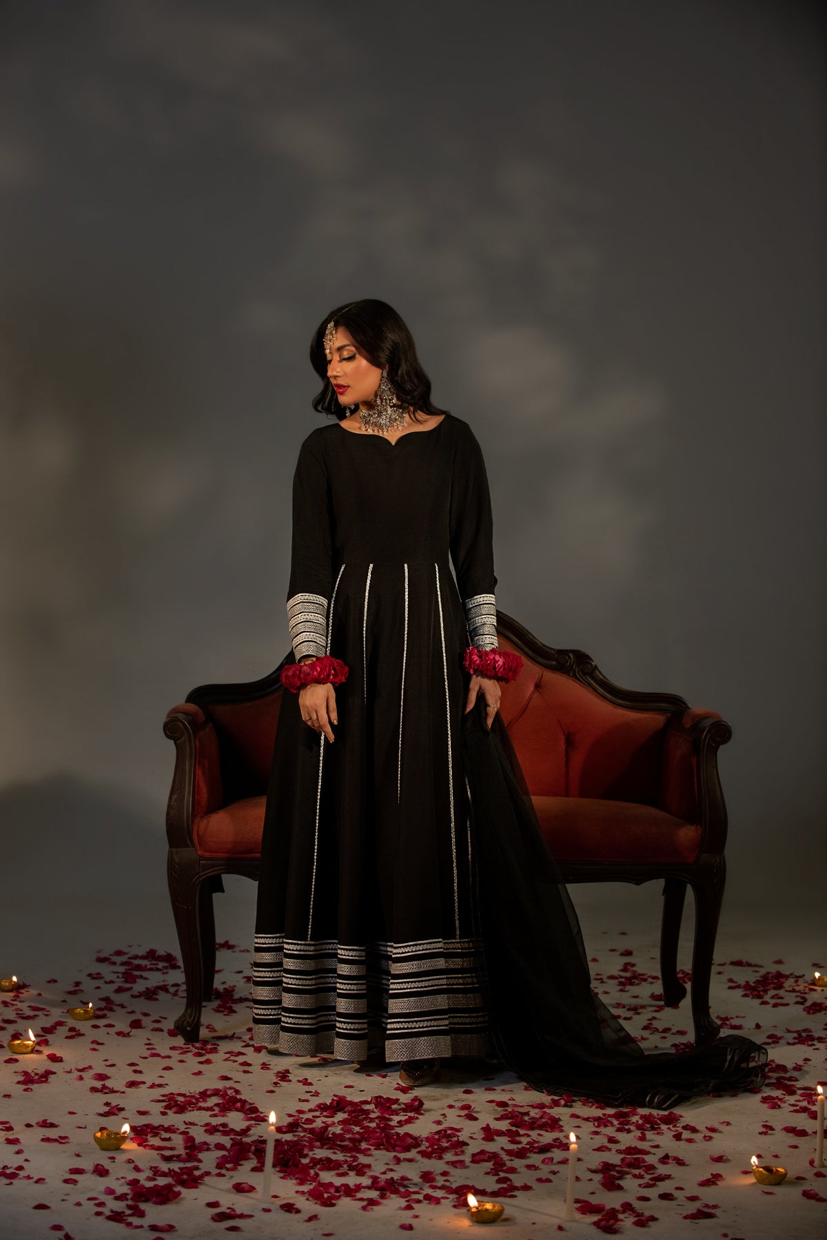 Woman in a black dress with red accents standing in front of a dark background with candles and rose petals.